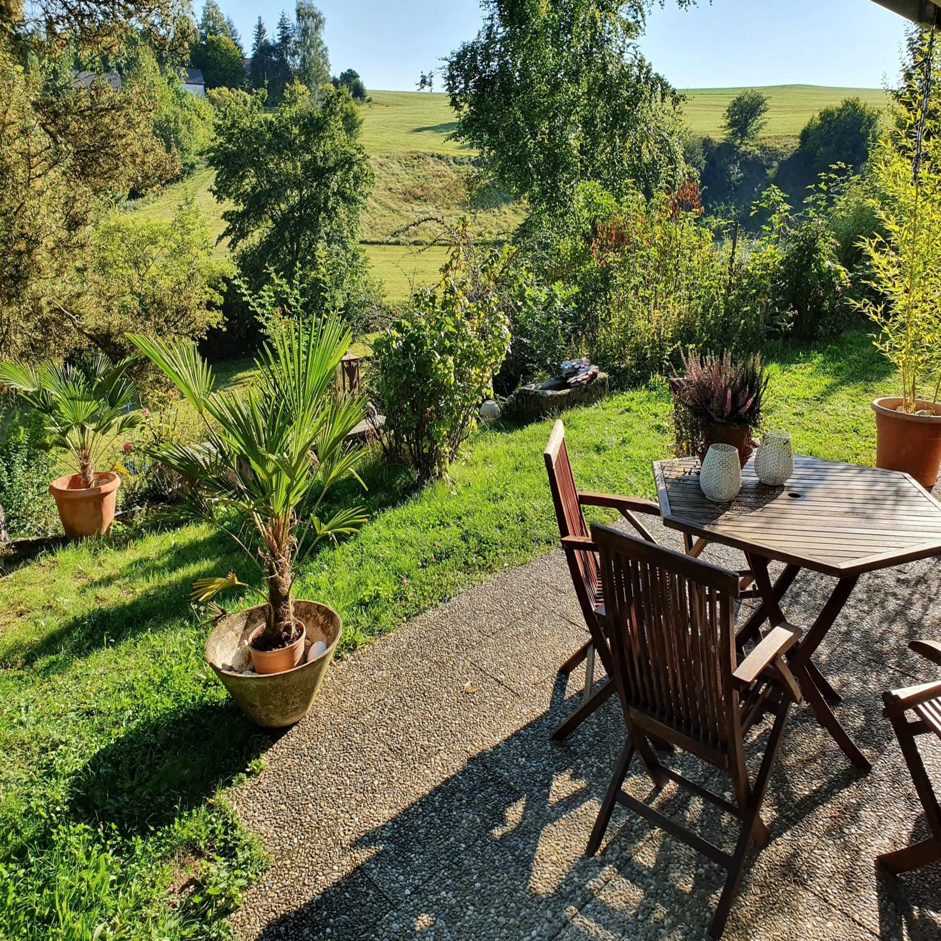 Terrasse mit Tisch und Stühlen in einem grünen Garten inmitten einer grünen Landschaft bei Sonnenschein.