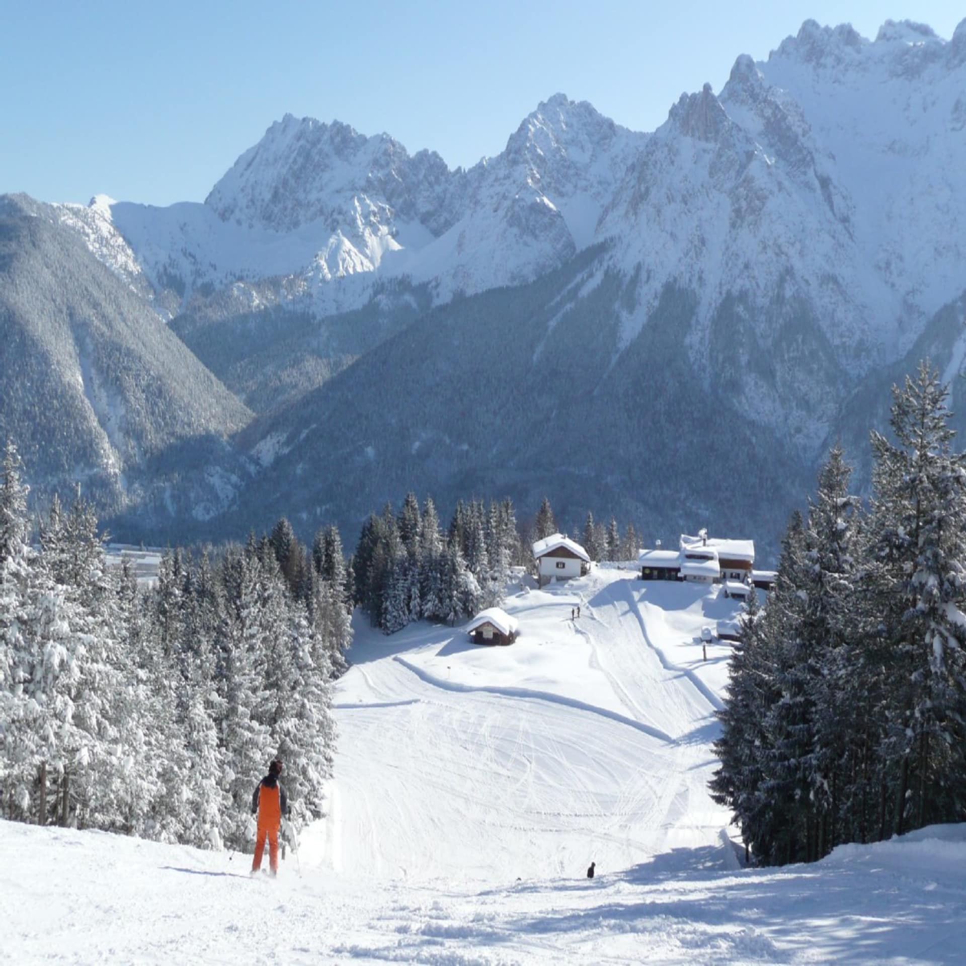 Eine Person in Orange auf einem Skihang, dahinter Skihütten. Die Sonne scheint.