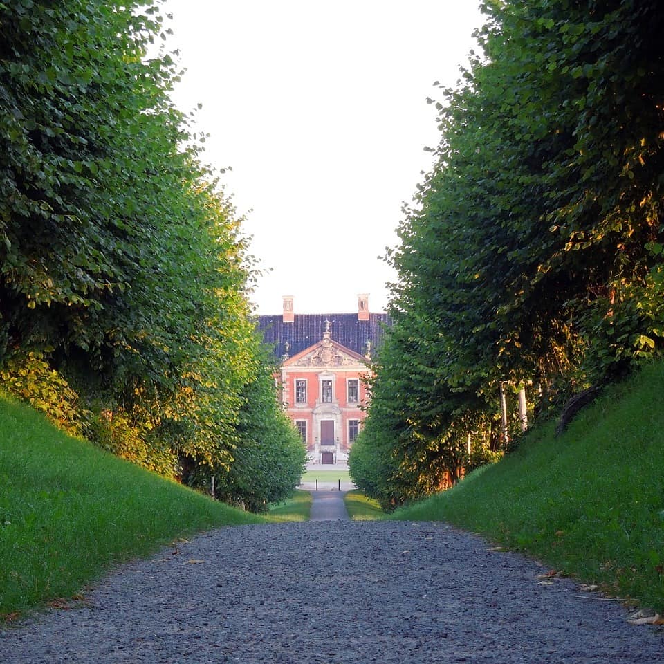 Ein herrschaftliches Gebäude thront am Ende einer Schotterallee die vom einer Hecke gesäumt ist.