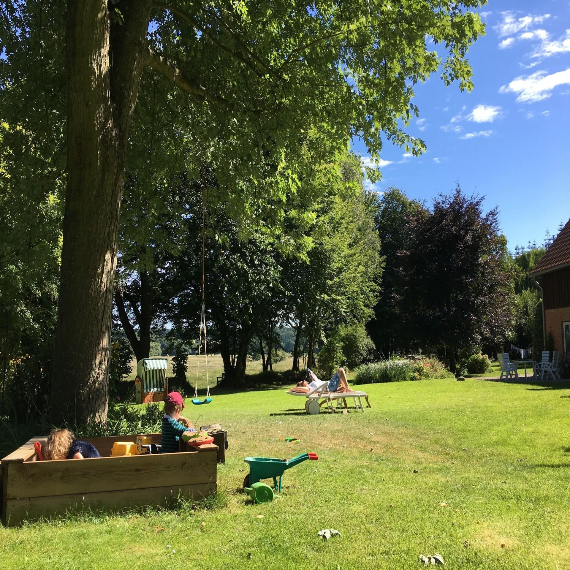 Garten: 2 Kinder spielen in Sandkiste, Frau liest ein Buch, ein Strandkorb, rechts ist ein Teil eines Bauernhauses zu sehen.