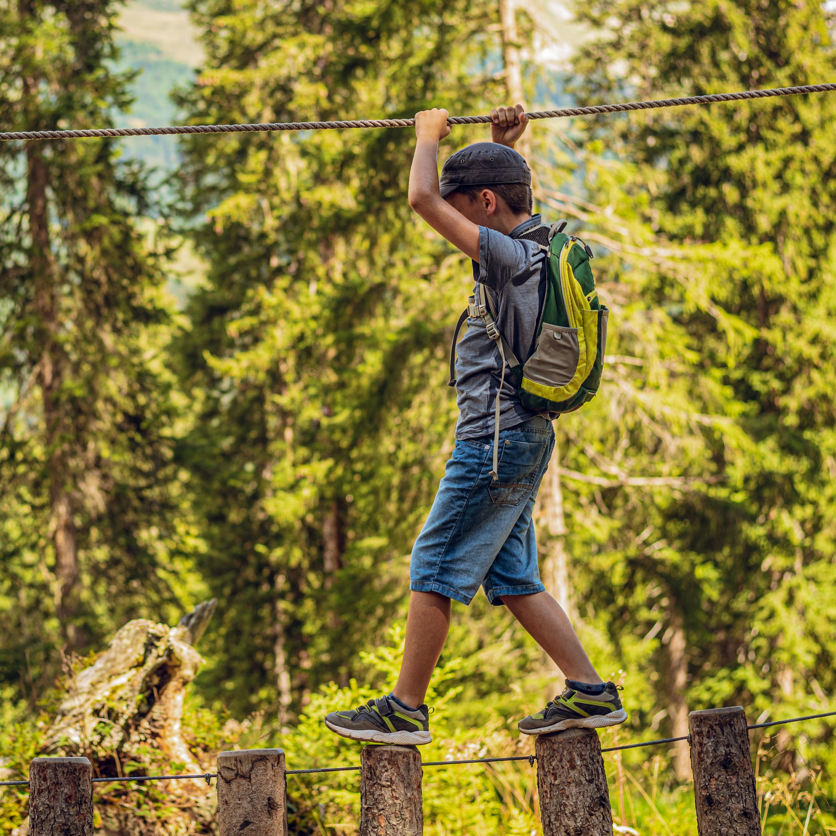 Junge in Sommerkleidung klettert über aufgereihte Baumstämme in einem Hochseilgarten.