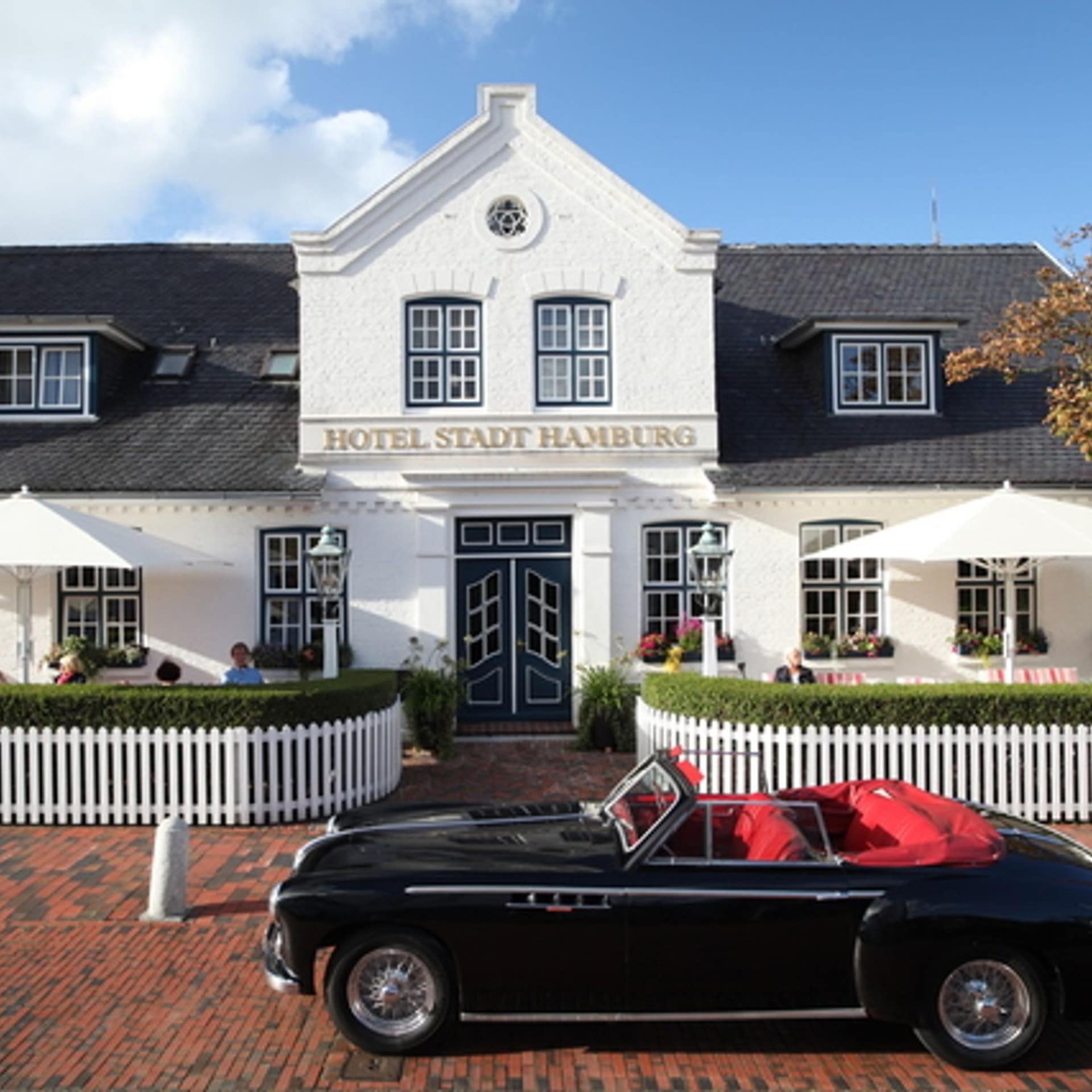 Ein schwarzer Oldtimer parkt vor einer weißen Villa auf Sylt. 