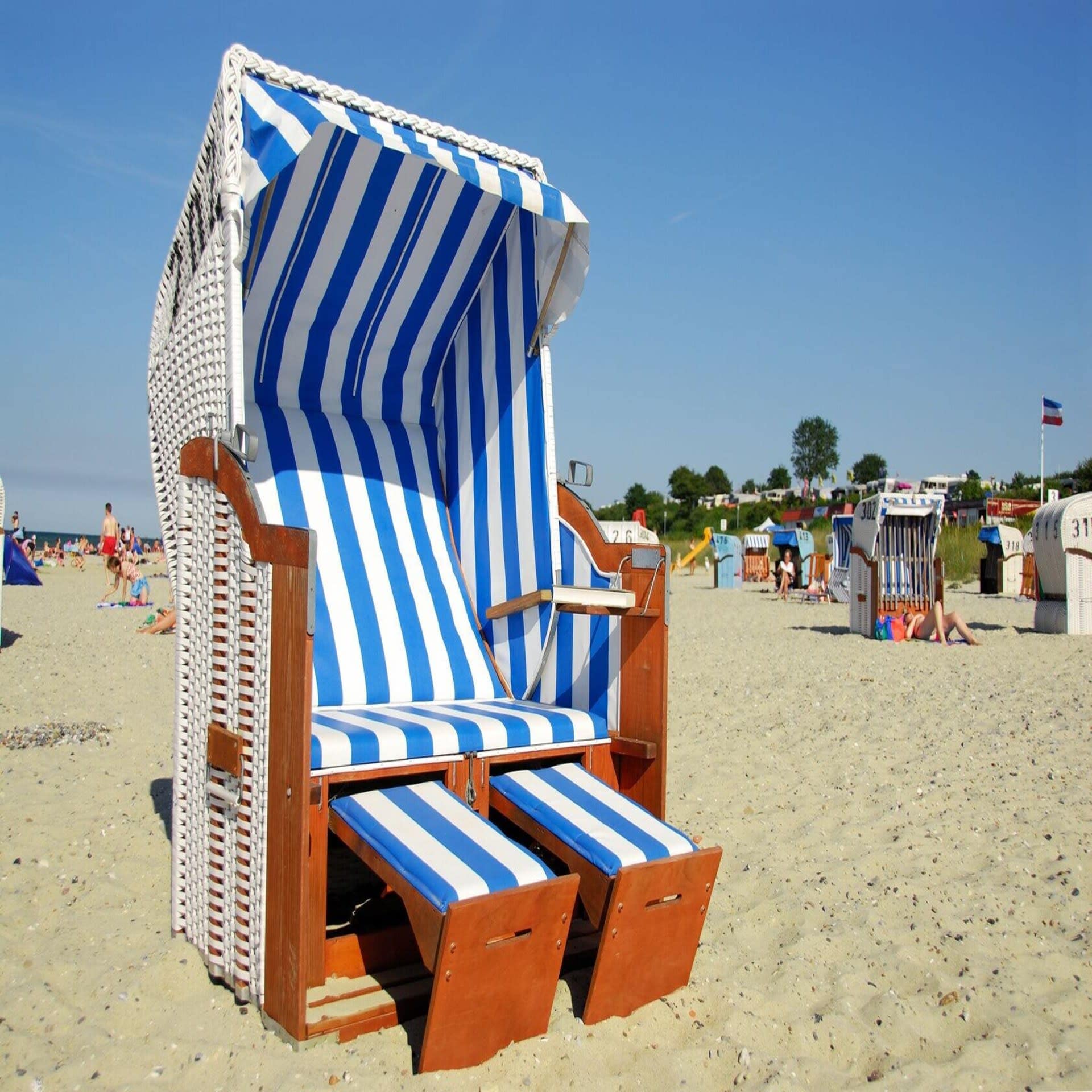 Ein blau-weiß gestreifter Strandkorb, dahinter Personen am Strand. Die Sonne scheint.