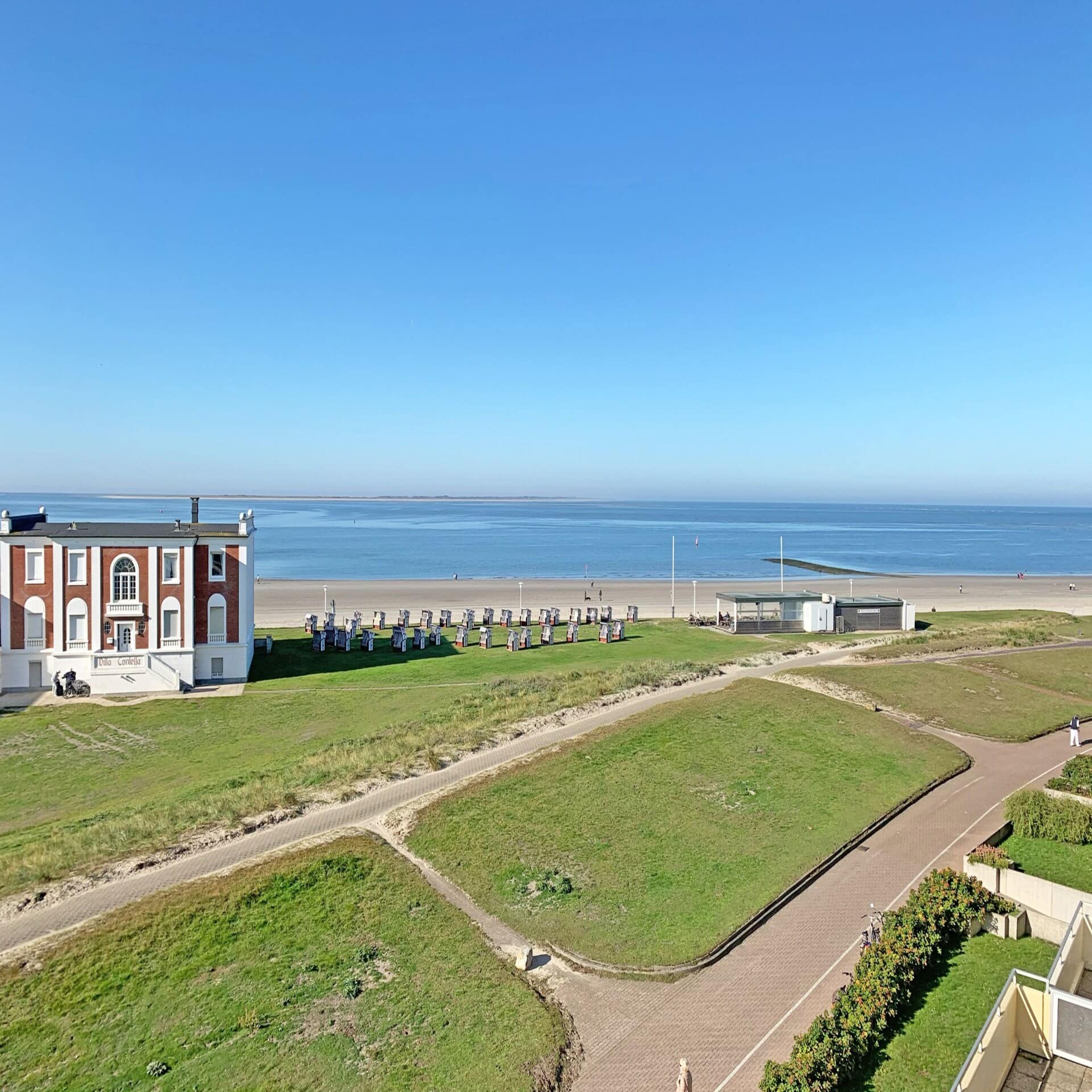 Blick vom Balkon über eine Wiese, auf den Strand und das Meer. Links ist ein rotes Friesenhaus zu sehen. 