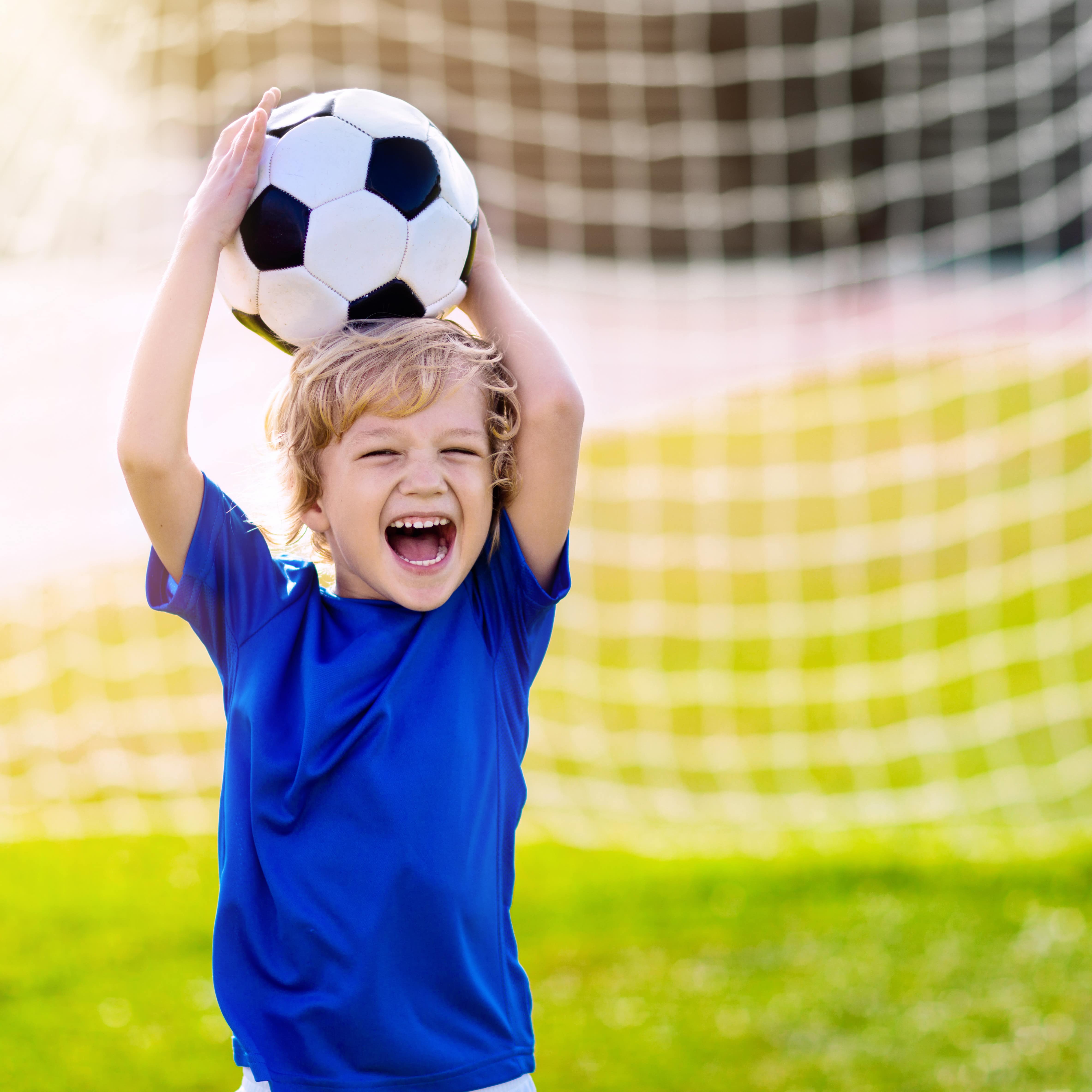 Ein kleiner Junge in blauem T-Shirt vor einem Fußballtor hält lachend einen Ball über dem Kopf.