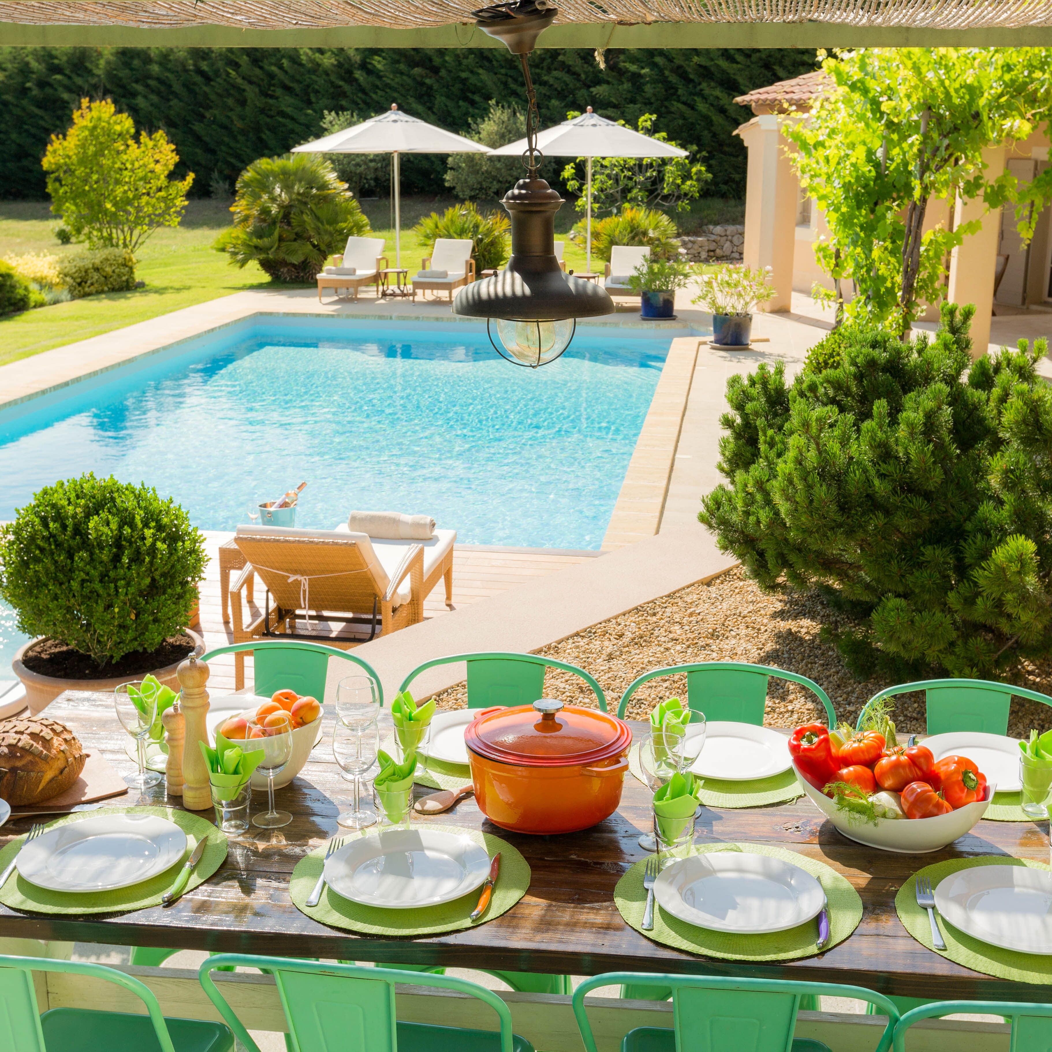 Hell gestrichenes Ferienhaus in der Provence mit großem Garten und Pool, neben dem im Schatten ein gedeckter Tisch wartet 