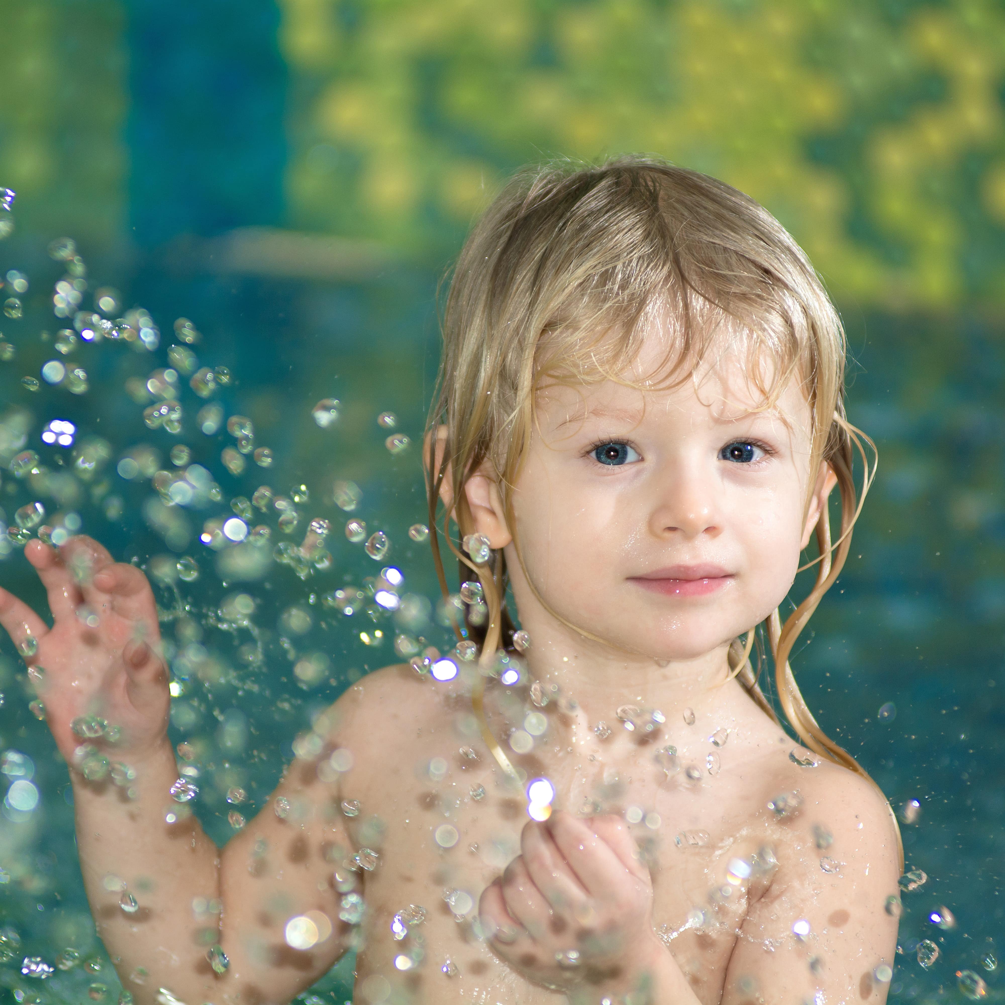 Kleinkind mit langen blonden Haaren im Wasser, davor spritzt glitzerndes Wasser.