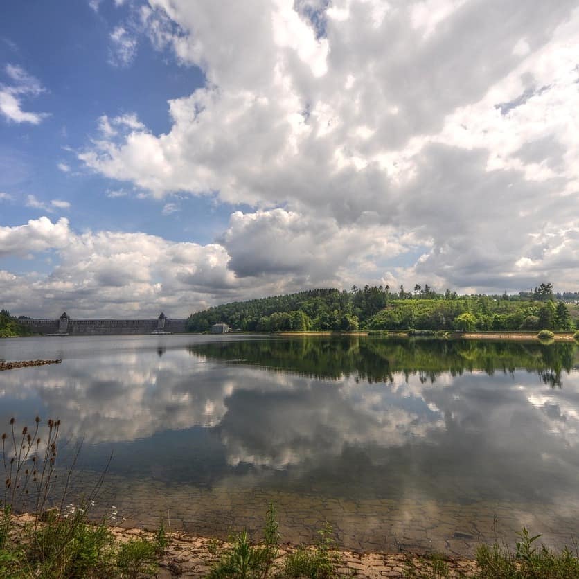 Blick über einen Stausee und die grüne Landschaft im Sauerland, im Wasser spiegeln sich die Wolken.