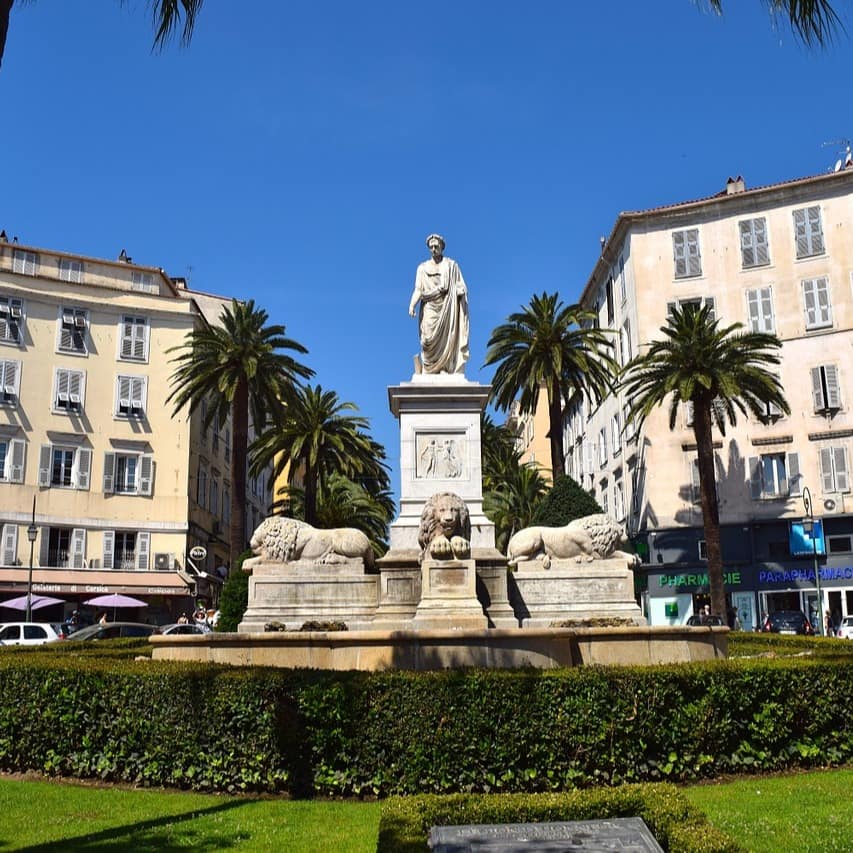 Blick auf ein Napoleon Denkmal in Ajaccio das von Palmen und Häusern gesäumt wird.