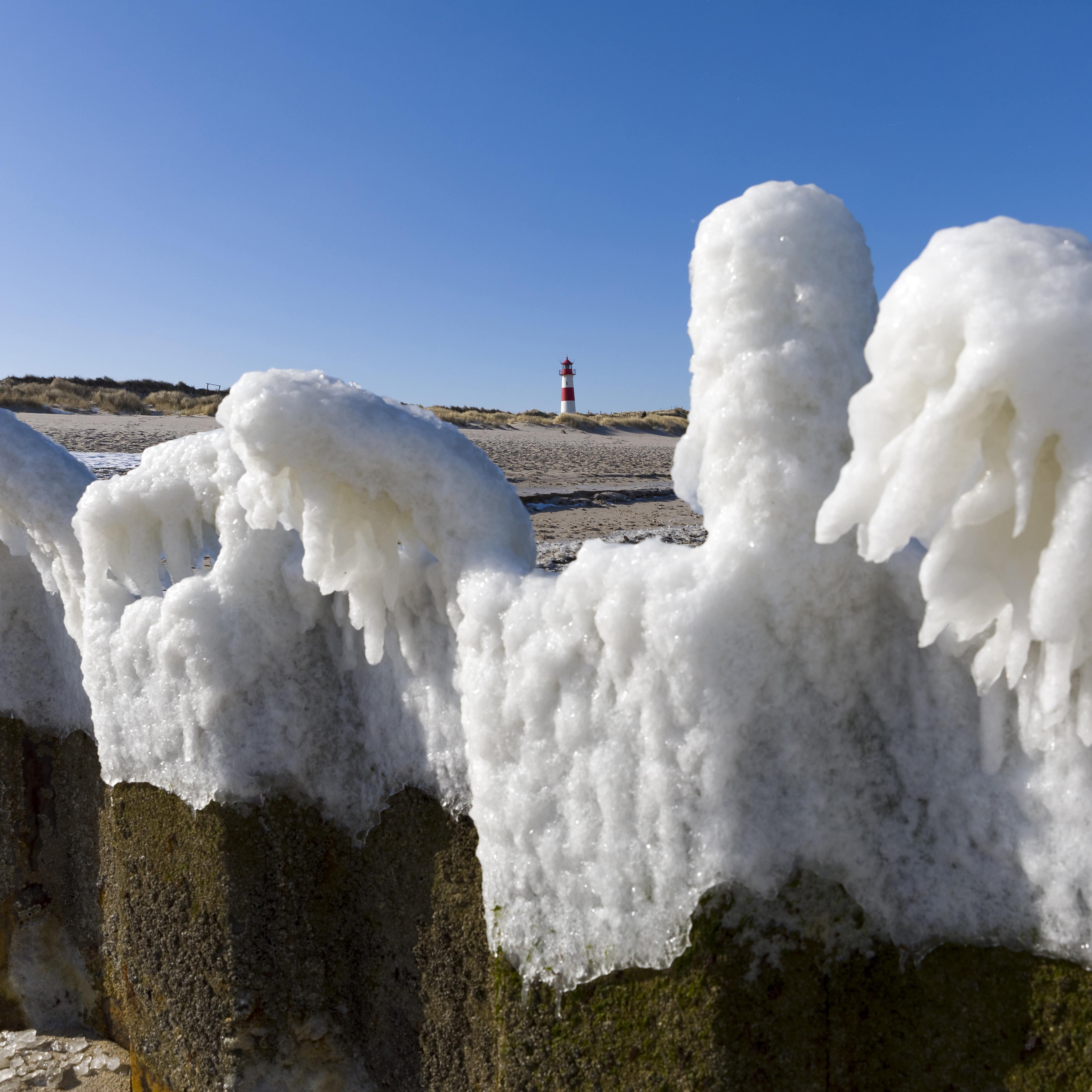 Vereiste Steinblöcke am Strand, im Hintergrund ein Leuchtturm.