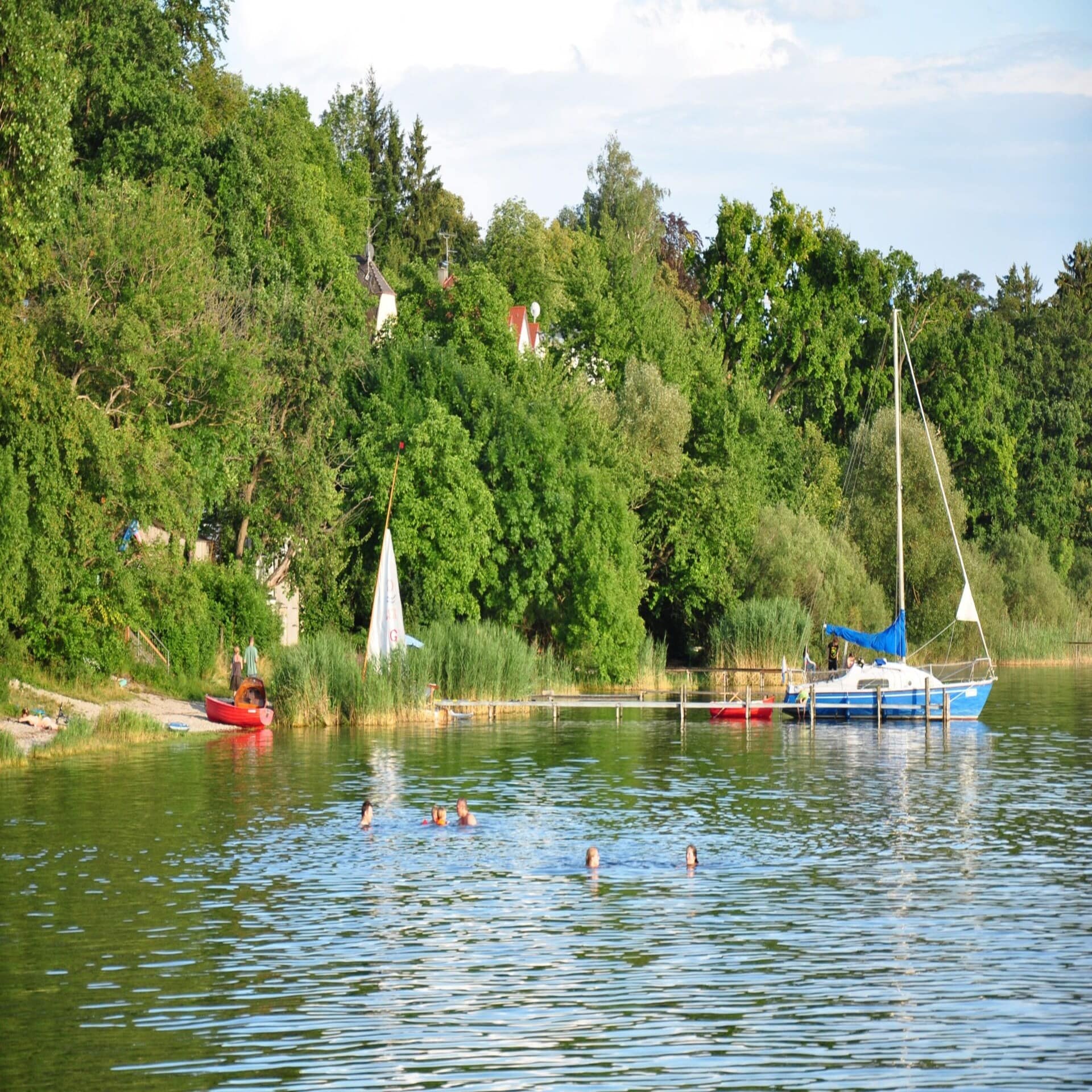 Leute schwimmen in einem See der von Bäumen umgeben ist. Ein kleines Segelboot ankert an einem Steg.  