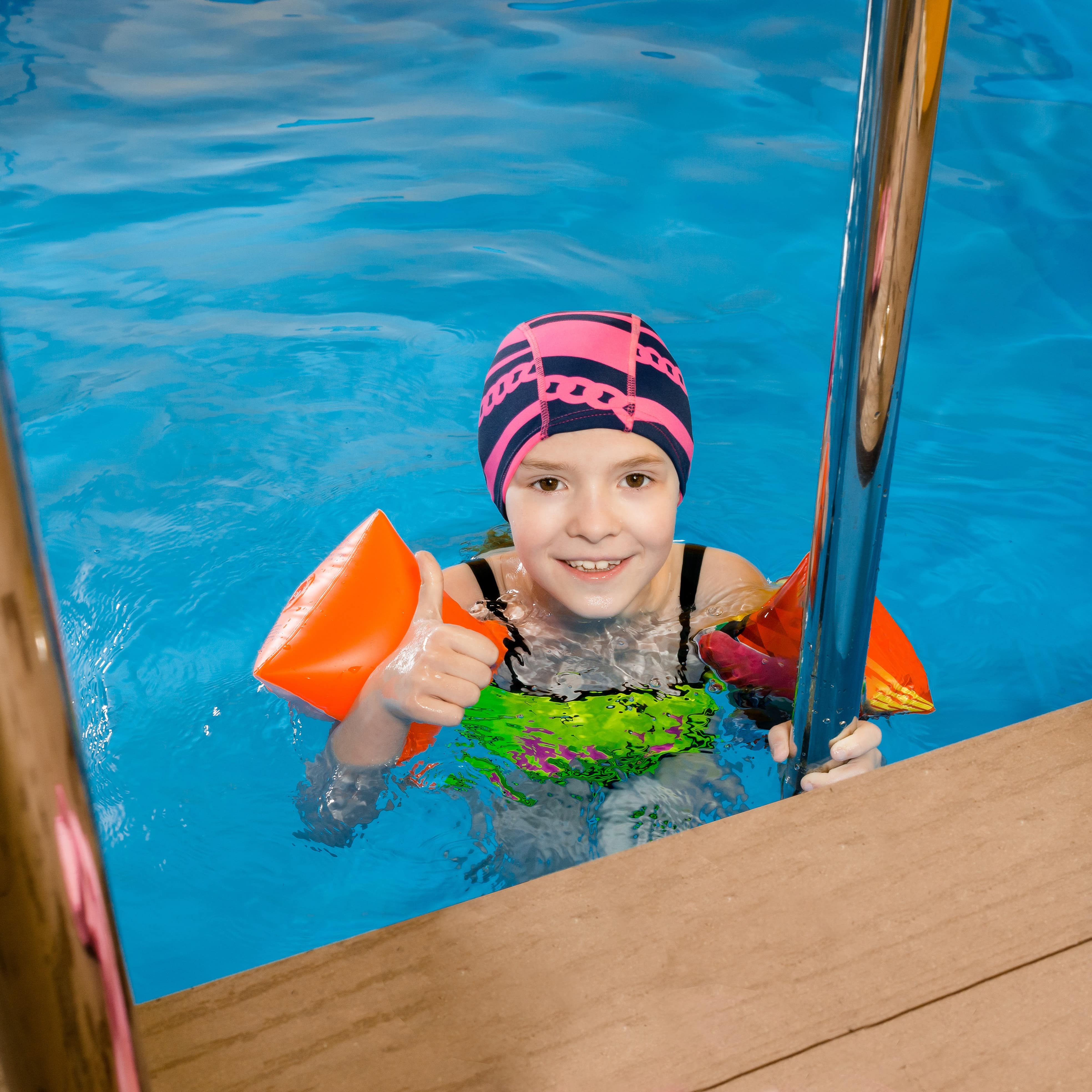 Ein Mädchen in einem Indoor-Pool hält sich an der Leiter fest. Sie hat eine Bademütze auf und Schwimmflügel an.