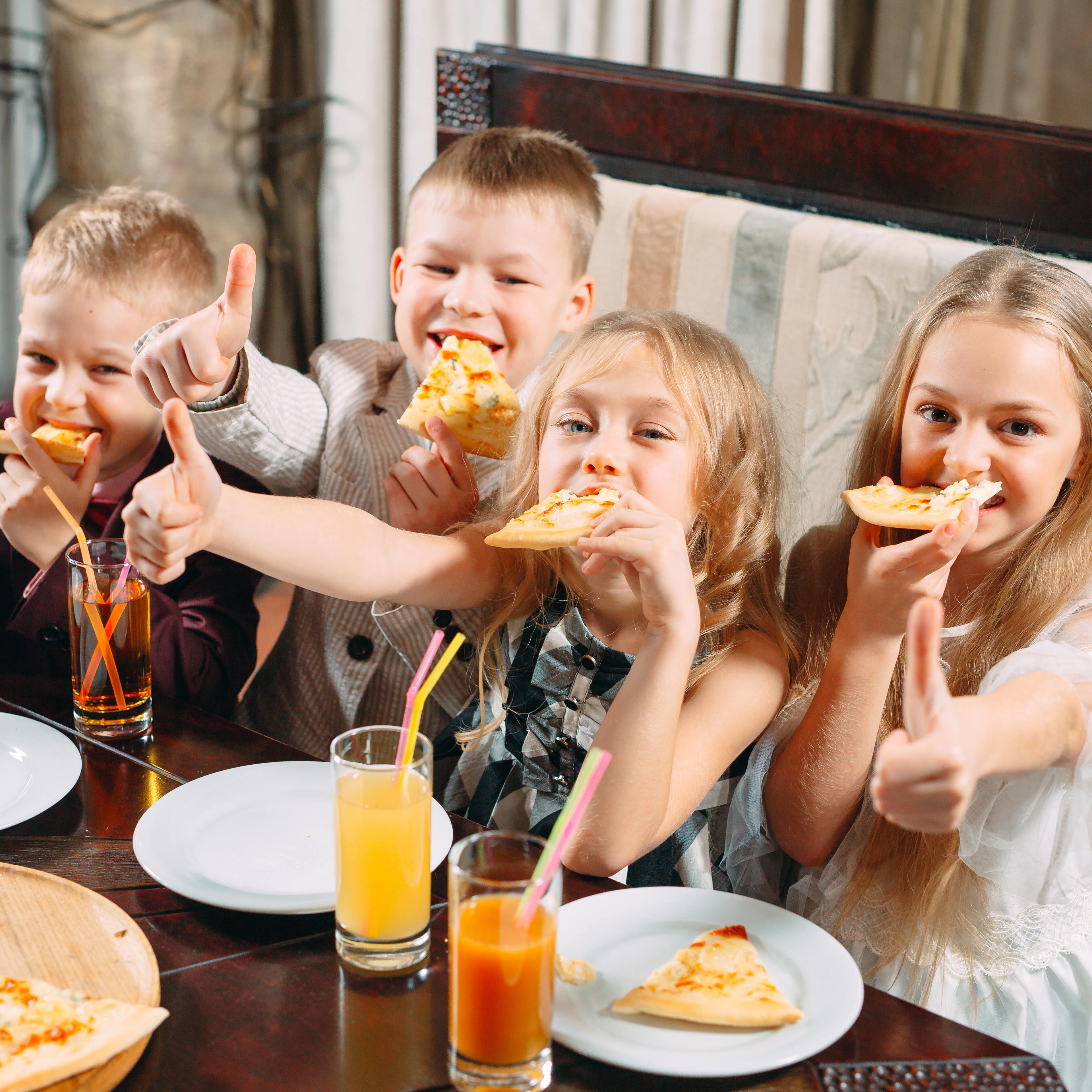 4 Kinder nebeneinander auf einer Bank im Restaurant, essen Pizza und trinken Saft. Alle haben den Daumen hochgestreckt.