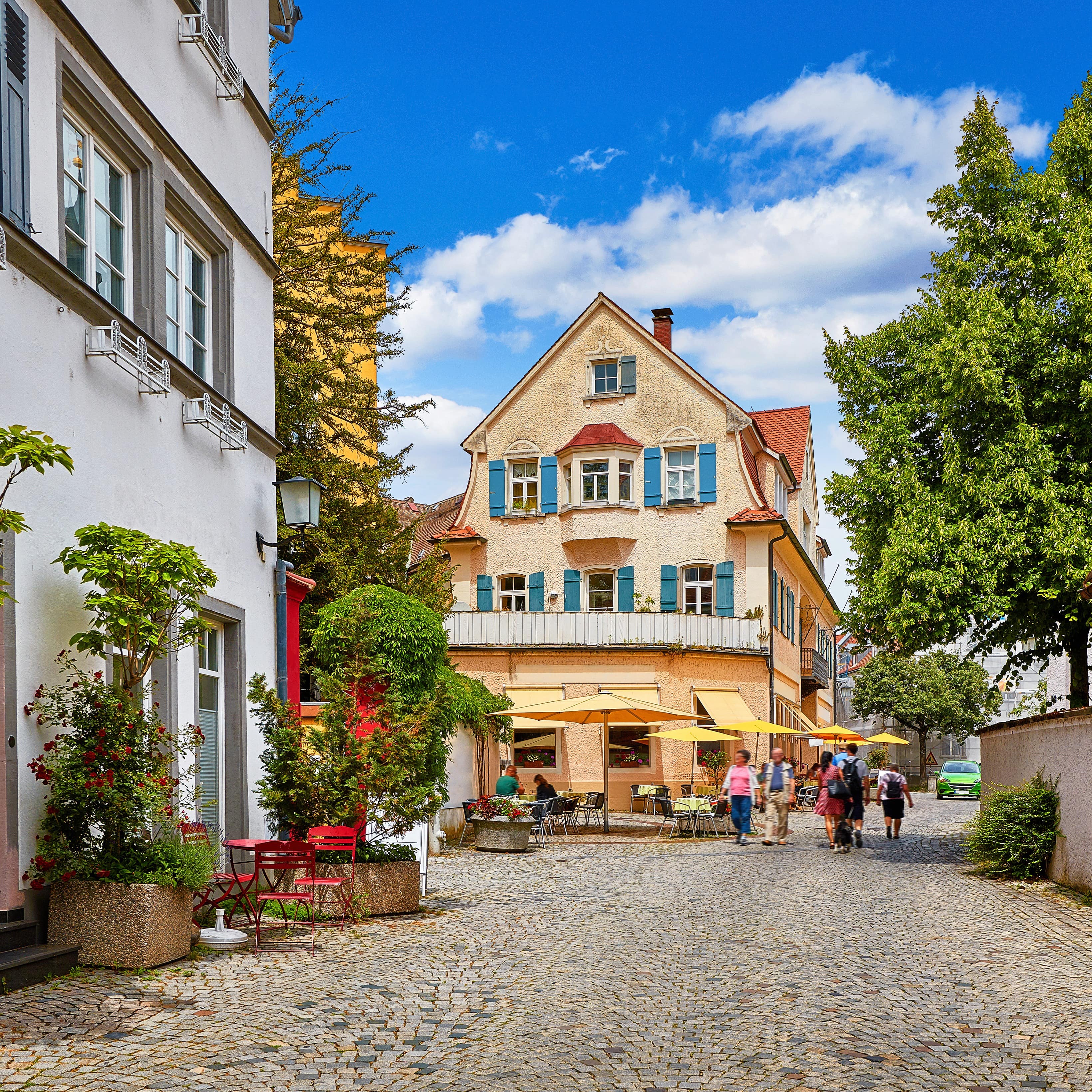 Ausschnitt einer Kopfsteingasse in Lindau mit älteren Häusern. Personen schlendern durch die Straße. 