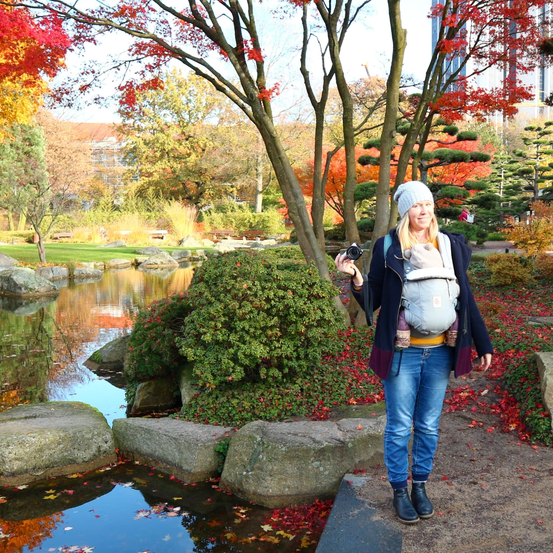 Frau mit Baby im Tragegurt und Kamera in der Hand im herbstlichen Park im Hamburg. 