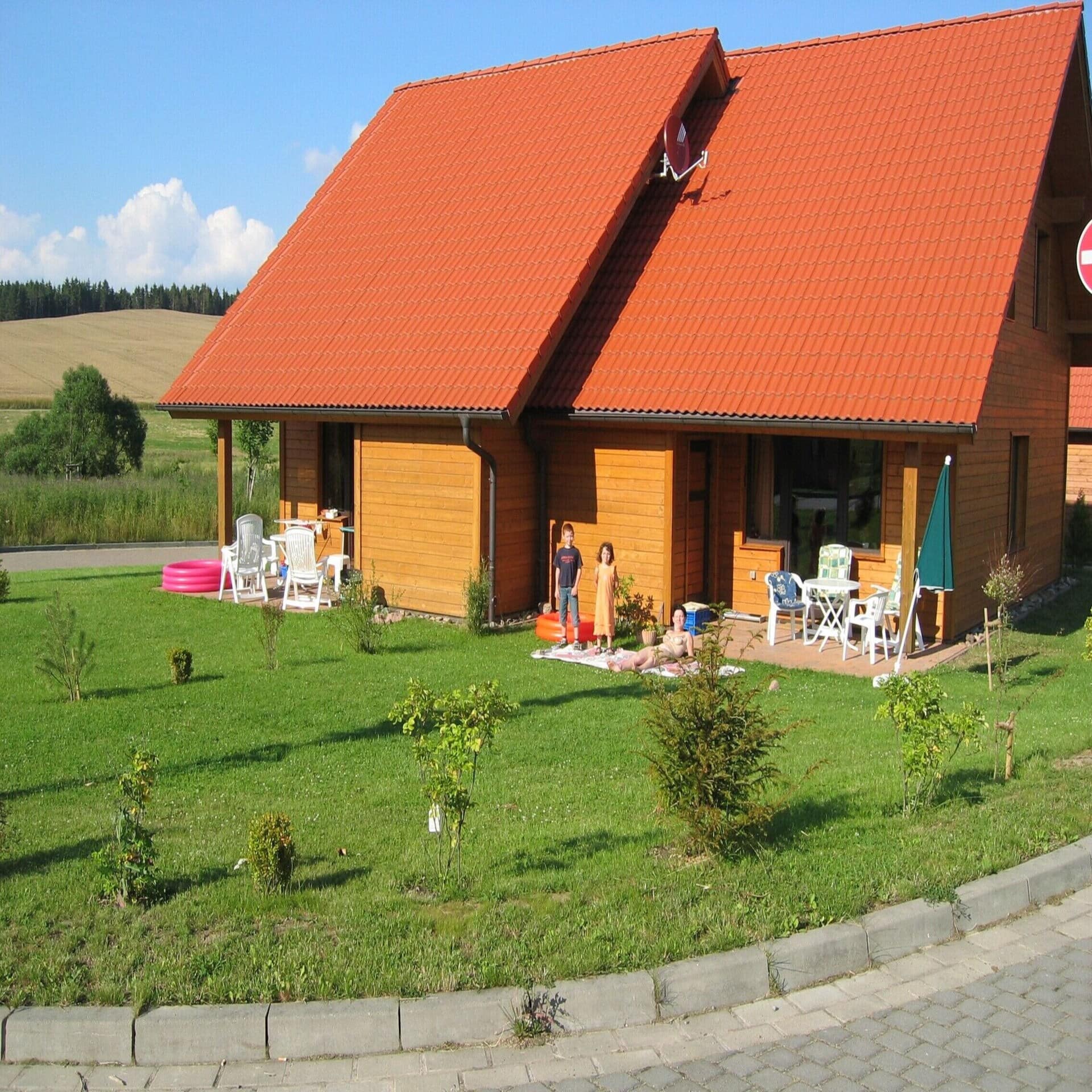 Holzhaus mit rotem Dach und Garten in dem ein Planschbecken, 2 Kinder und 1 Frau sind.