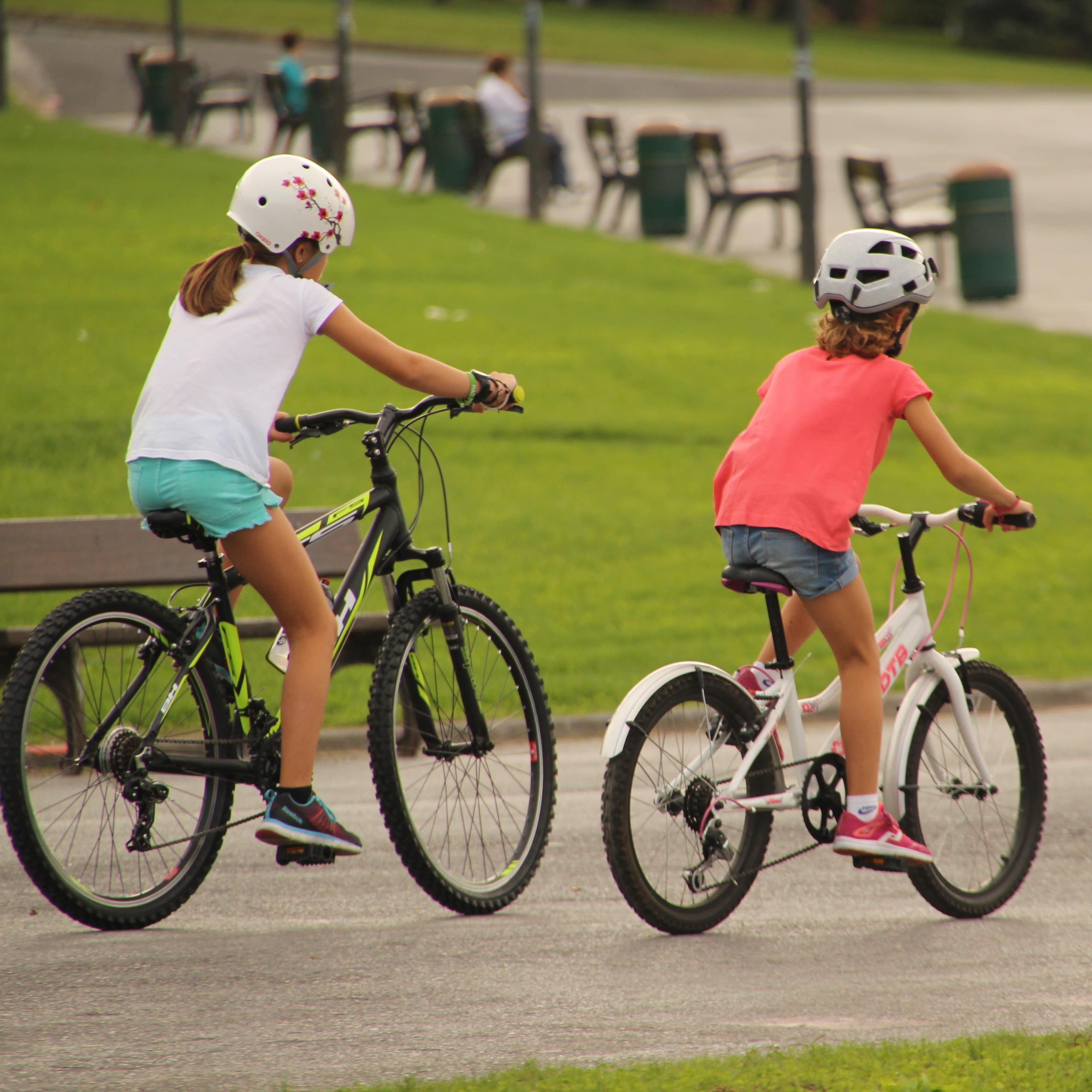 2 Mädchen in Sommerkleidung fahren mit Fahrrädern durch einen Park. Die Mädchen haben Helme auf. 