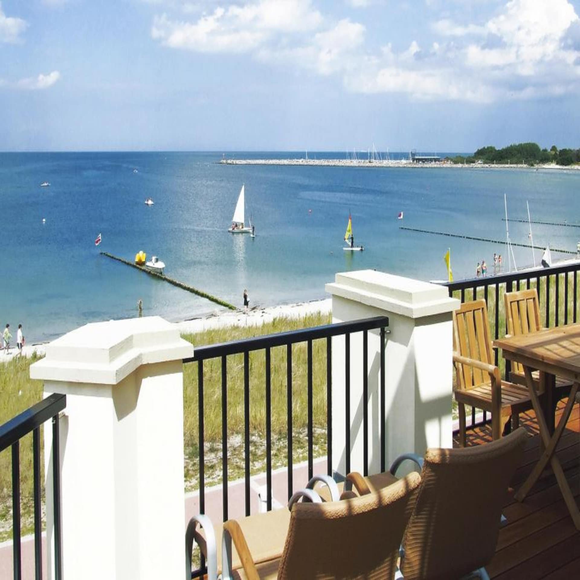 Blick von einem Balkon auf den Strand und die Ostsee. Im Wasser sind Surfer und kleine Segelboote. 