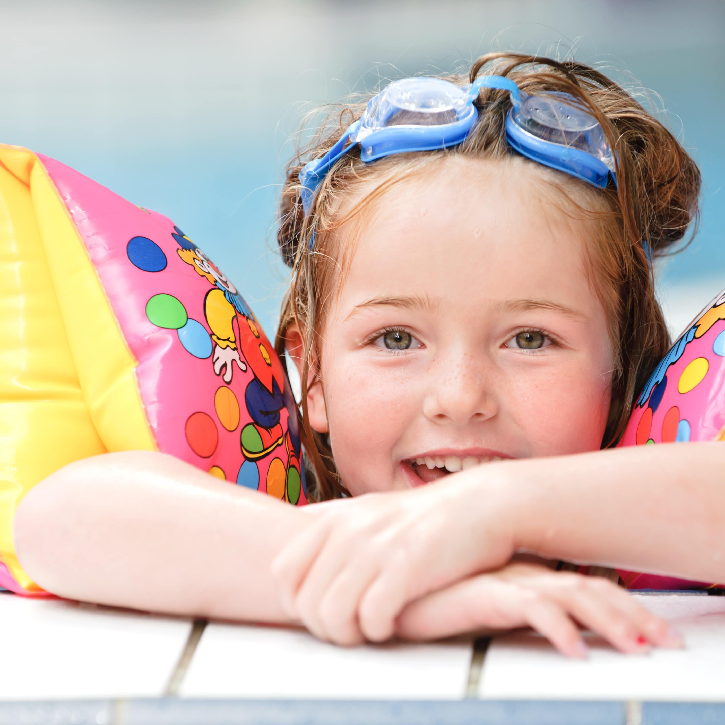 Nahaufnahme: Ein Mädchen mit bunten Schwimmflügeln und Schwimmbrille am Beckenrand im Wasser.