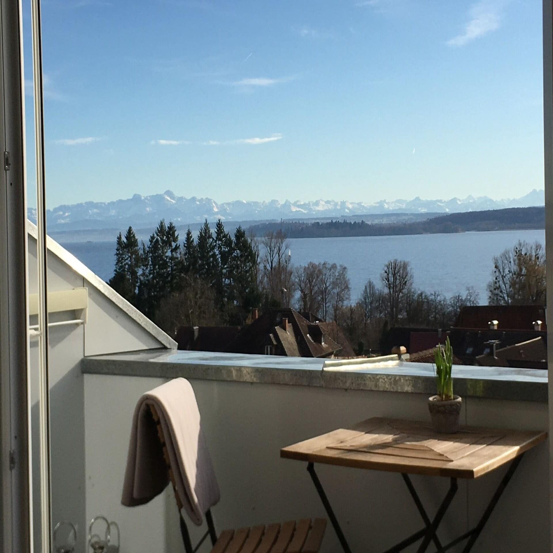 Balkon mit Tisch und Stühle und Blick über Häuser und Bäume hinweg auf den Bodensee. Im Hintergrund Berge.