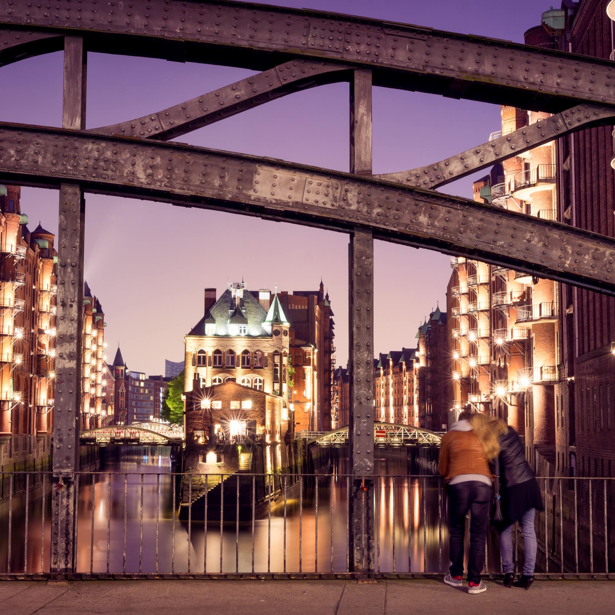 Rückenansicht: Ein Paar steht abends auf einer Brücke und schaut auf die hell erleuchtete Speicherstadt.