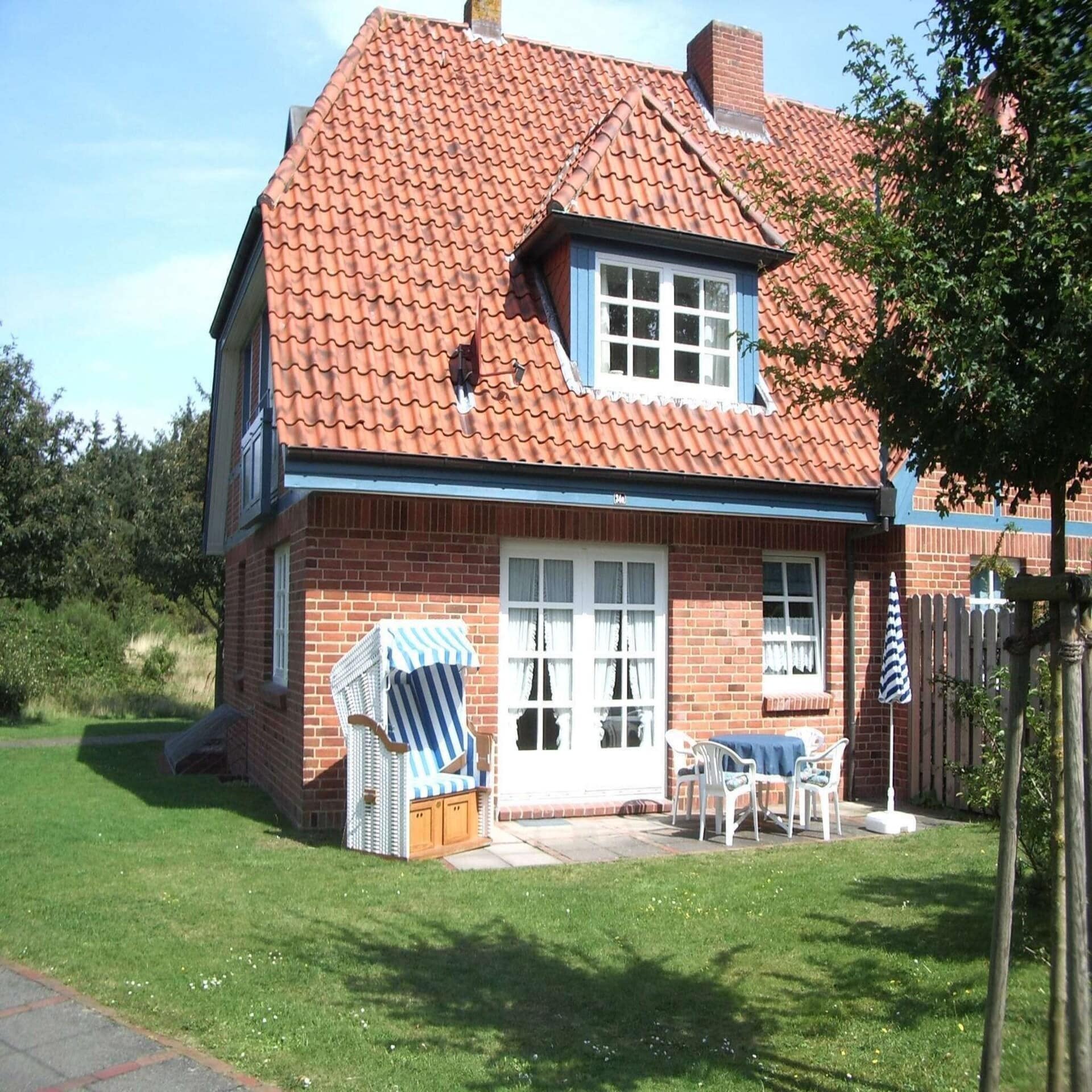  Rotes Friesenhaus mit weißen Fenstern. Blau-weiß gestreifter Strandkorb und Gartenmöbel stehen auf der Terrasse.