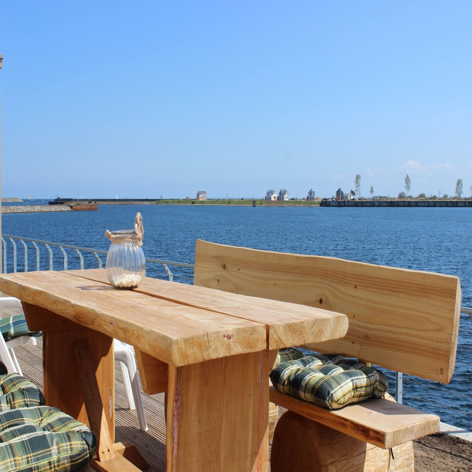 Terrasse mit Holztisch und Sitzbänken direkt am Wasser, im Hintergrund einige Häuser.