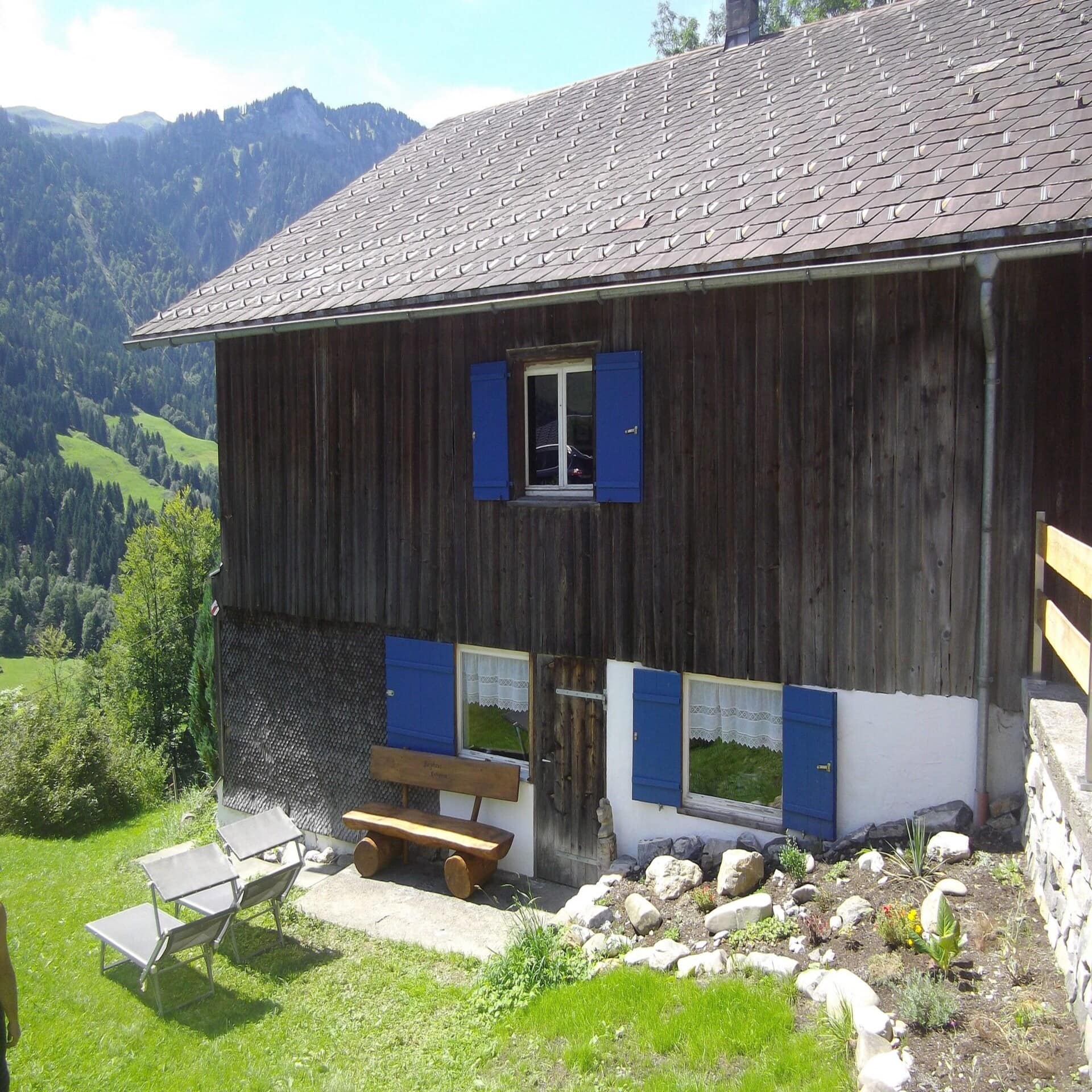 Braunes Bauernhaus mit blauen Fensterläden. Davor eine Holzbank und 2 Sonnenliegen.