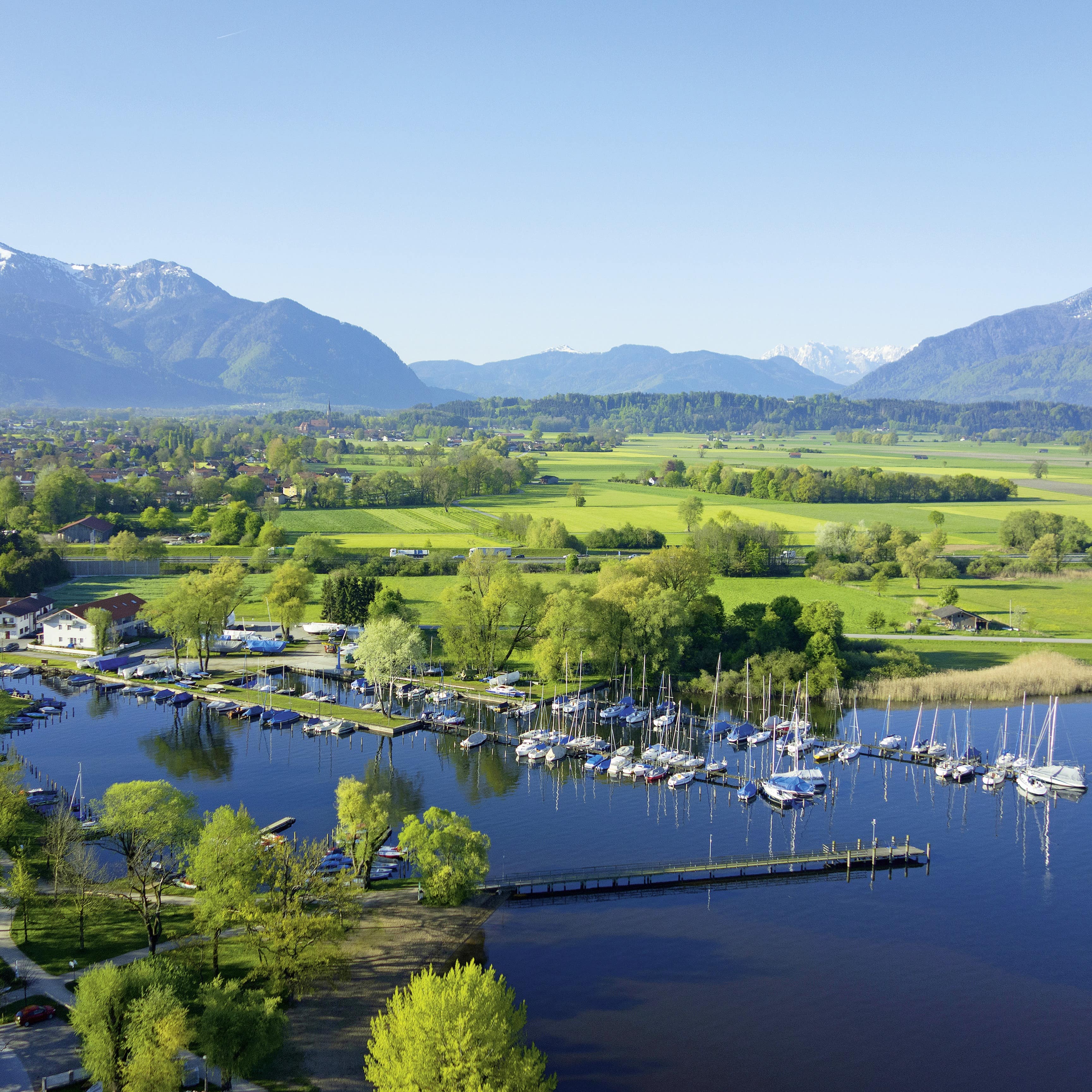 Panoramablick: Ein kleiner Hafen mit Segelbooten am Chiemsee, dahinter viel Grün und die Alpen.