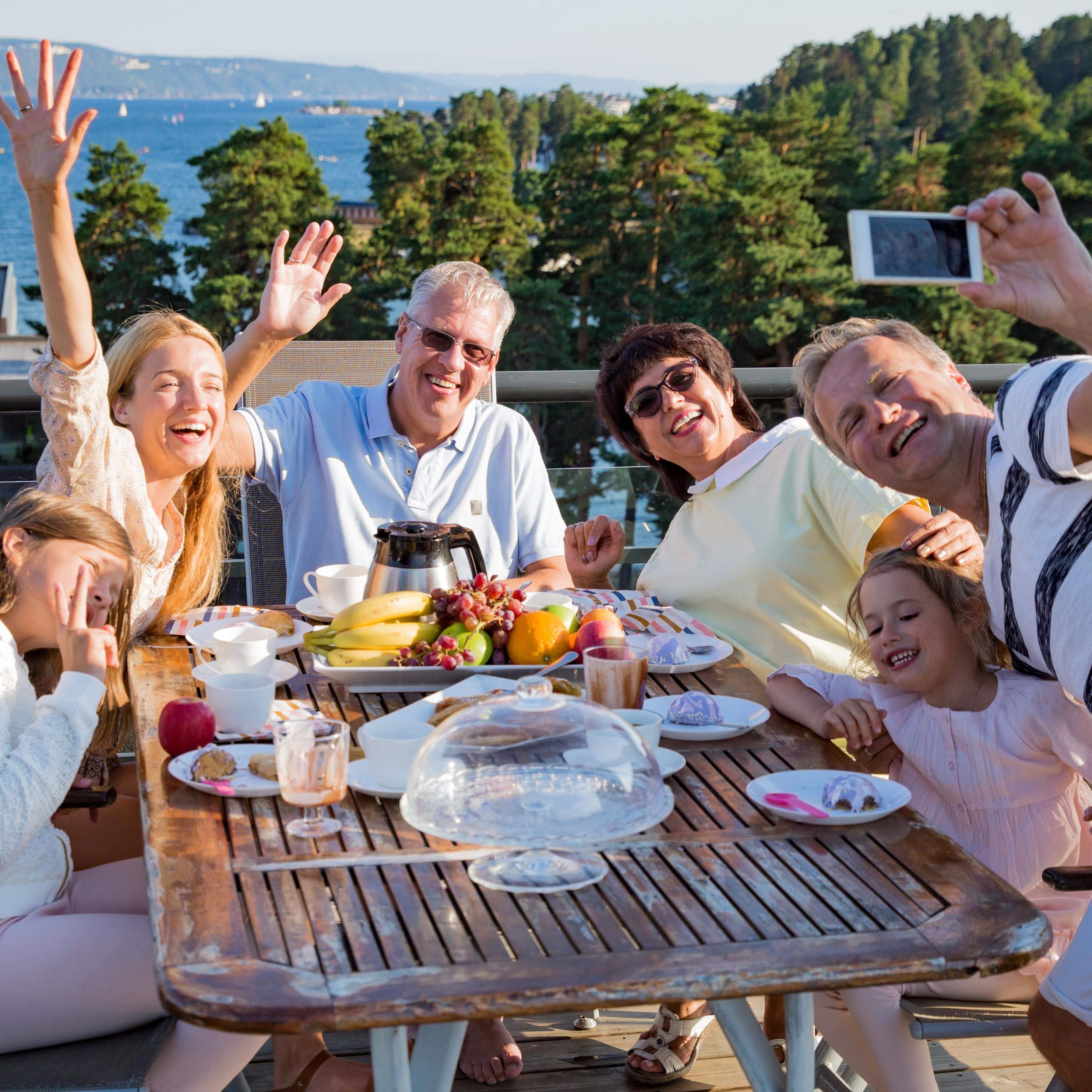 Familie mit Kindern und Großeltern am Frühstückstisch auf einer Terrasse machen ein Foto. Im Hintergrund ein See.