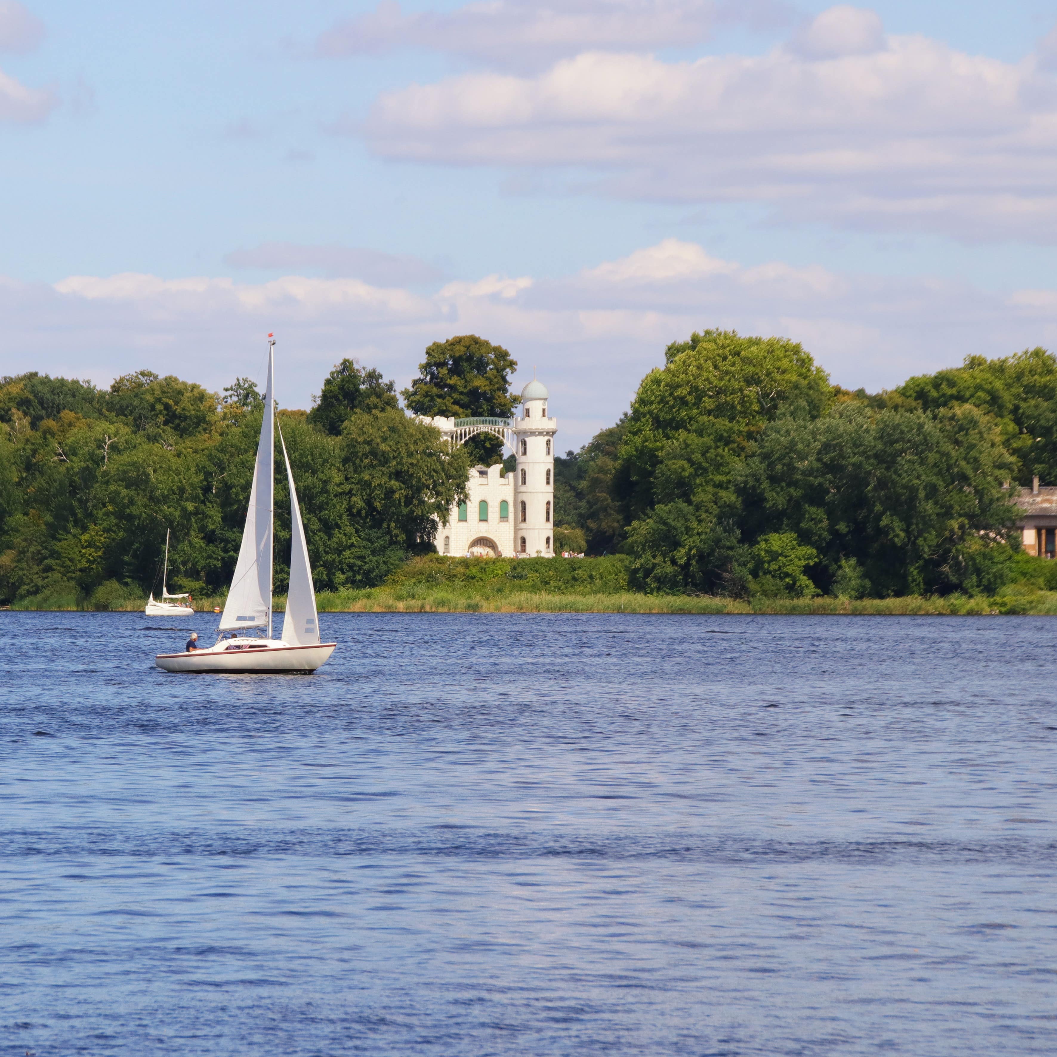 Blick über das Wasser, auf dem ein Segelboot schippert, dahinter die Pfaueninsel mit Schloss und Bäumen.