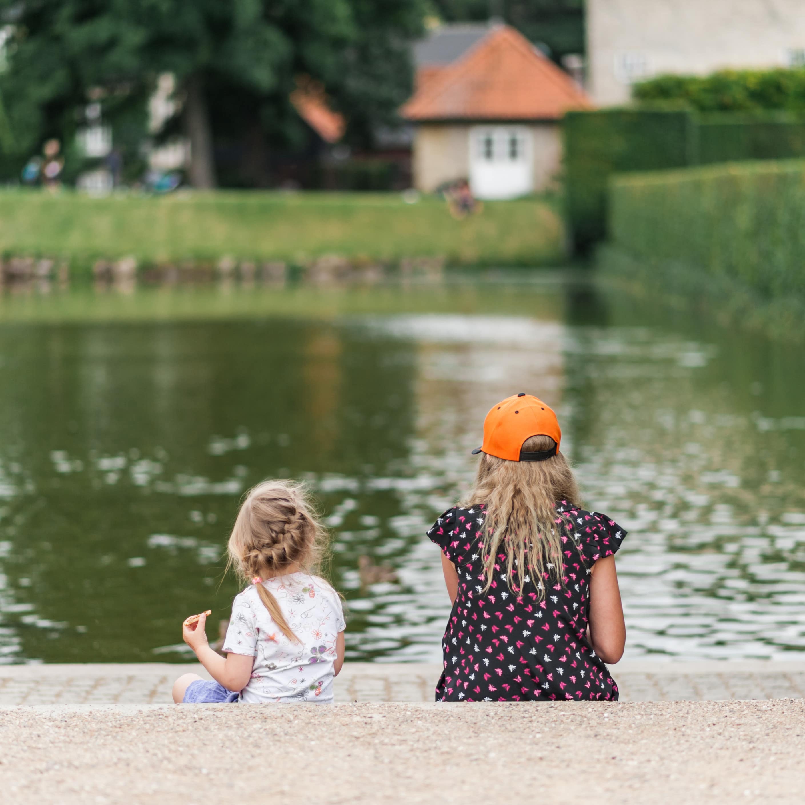 Rückenansicht: 2 blonde Mädchen in Sommerkleidungsitzen am Seeufer.