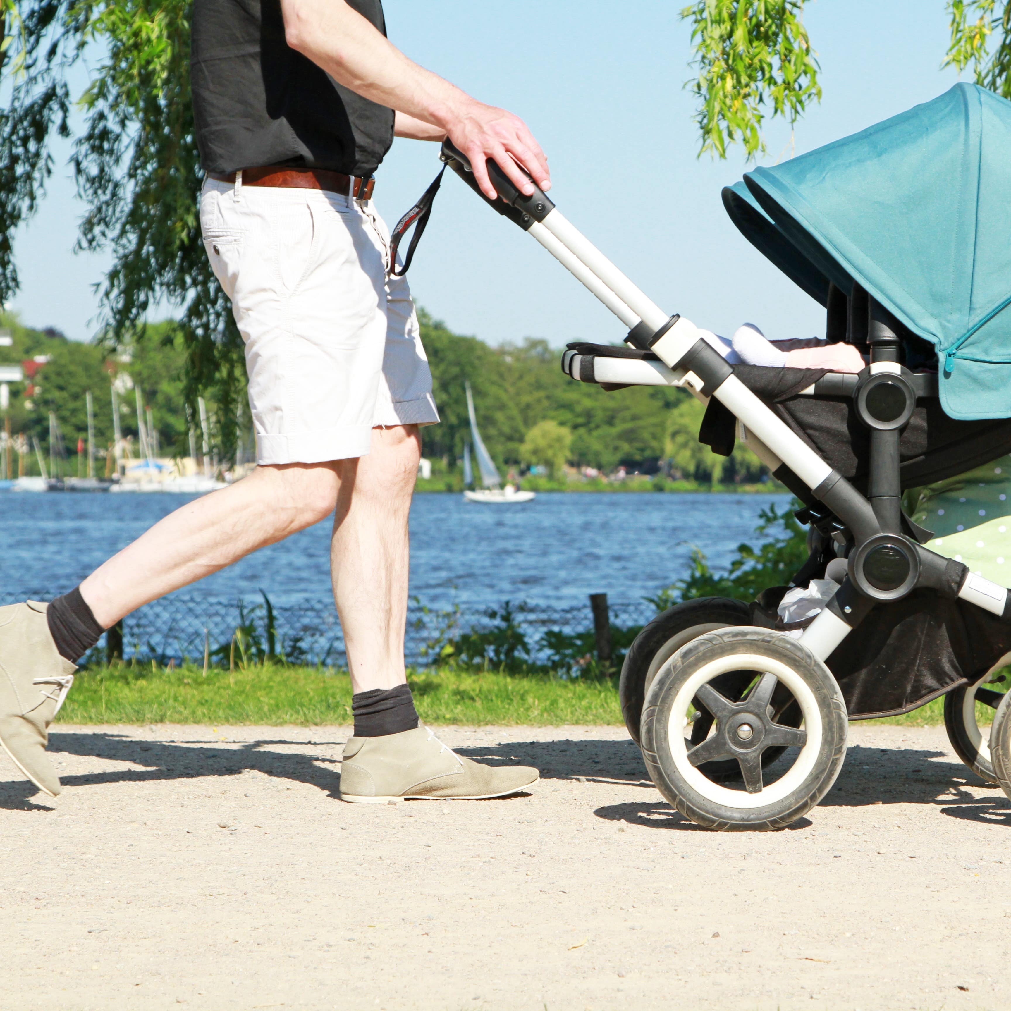 Nahaufnahme: Ein Mann in Sommerkleidung schiebt einen Kinderwagen am Flussufer. 