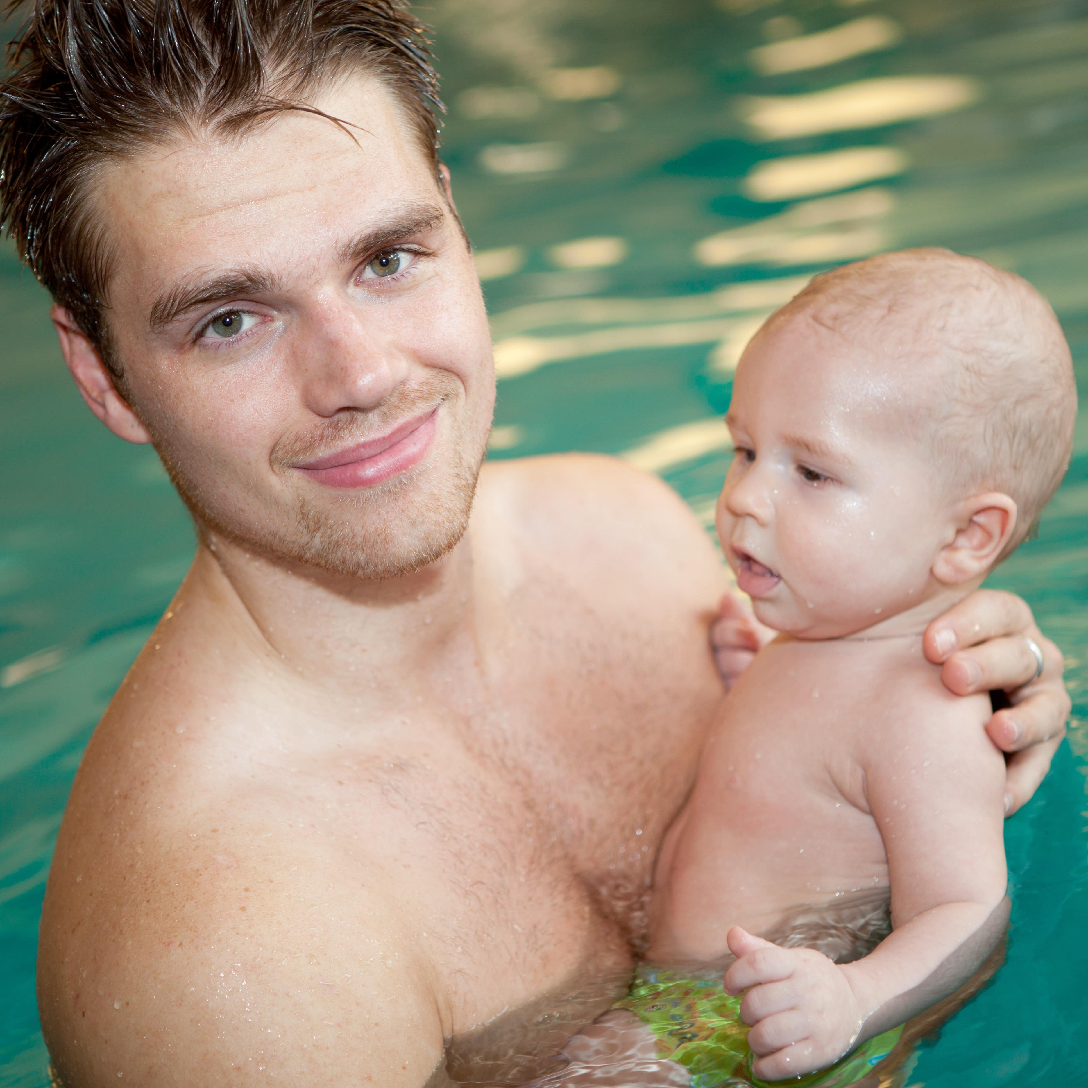 Vater mit Baby auf dem Arm im Wasser lächelt in die Kamera.
