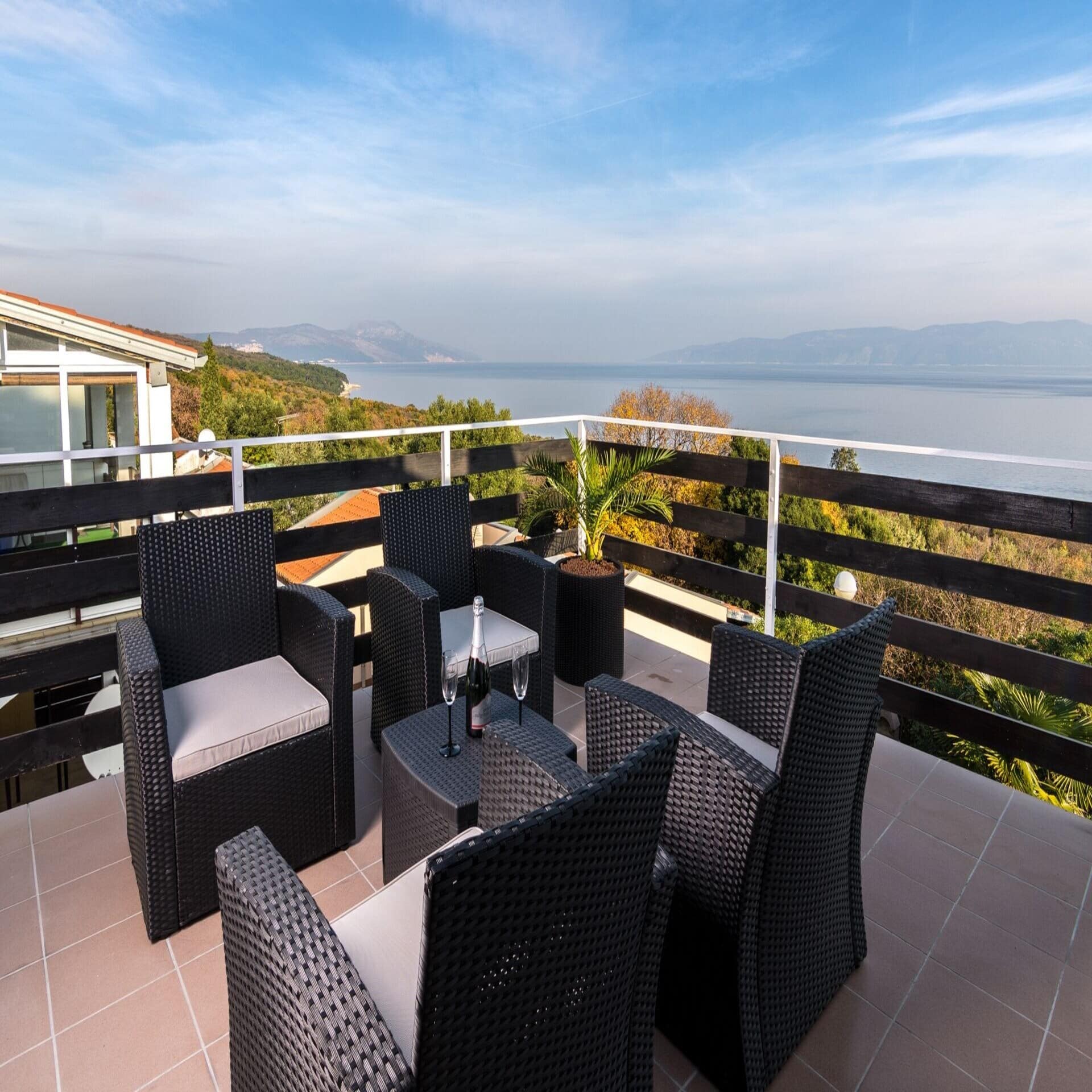 Balkon mit 4 dunklen Korbsesseln und Tisch und Blick auf ein Haus, das grüne Umland und das Meer. 