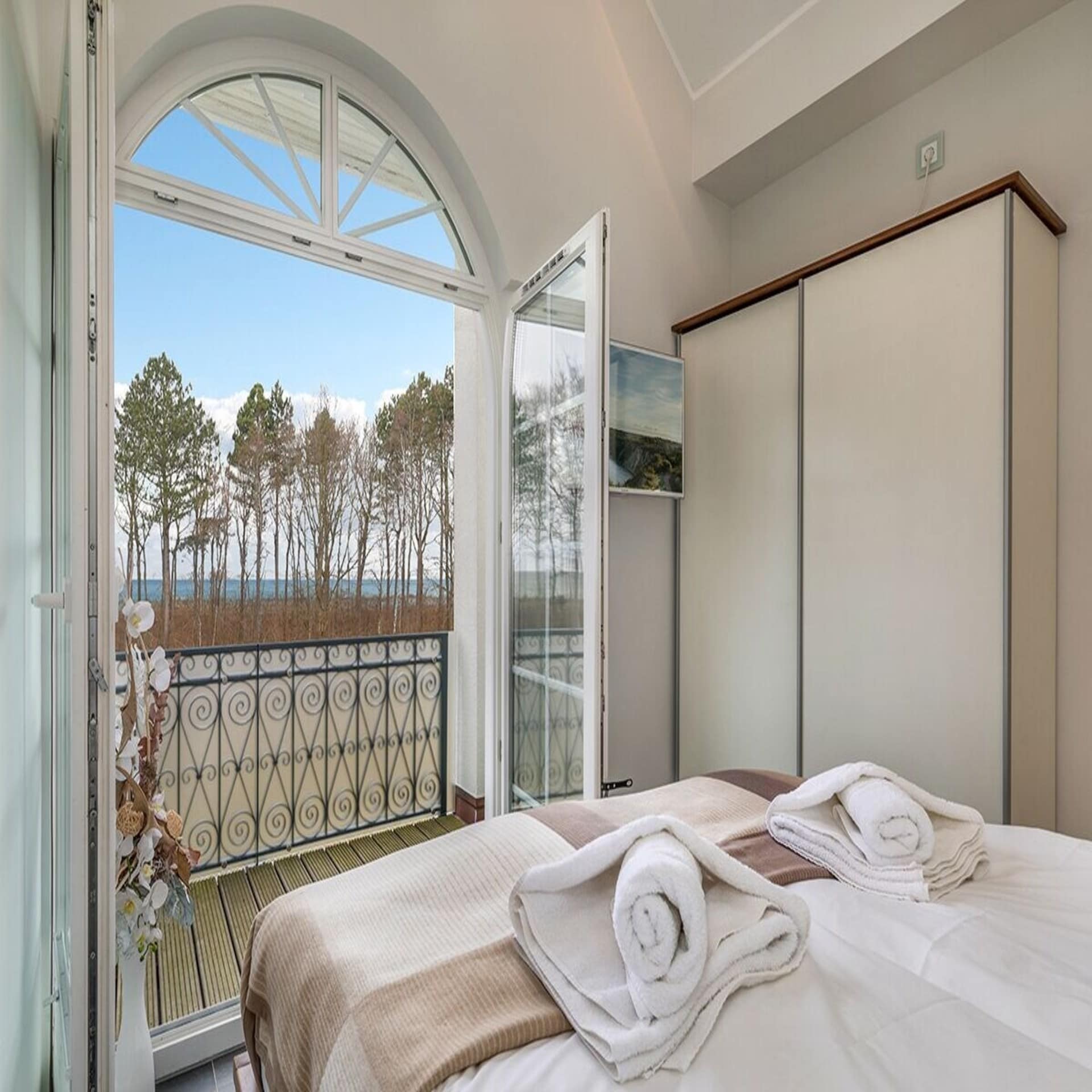Schlafzimmer mit geöffneter Balkontür mit Blick auf das Meer und Bäume.
