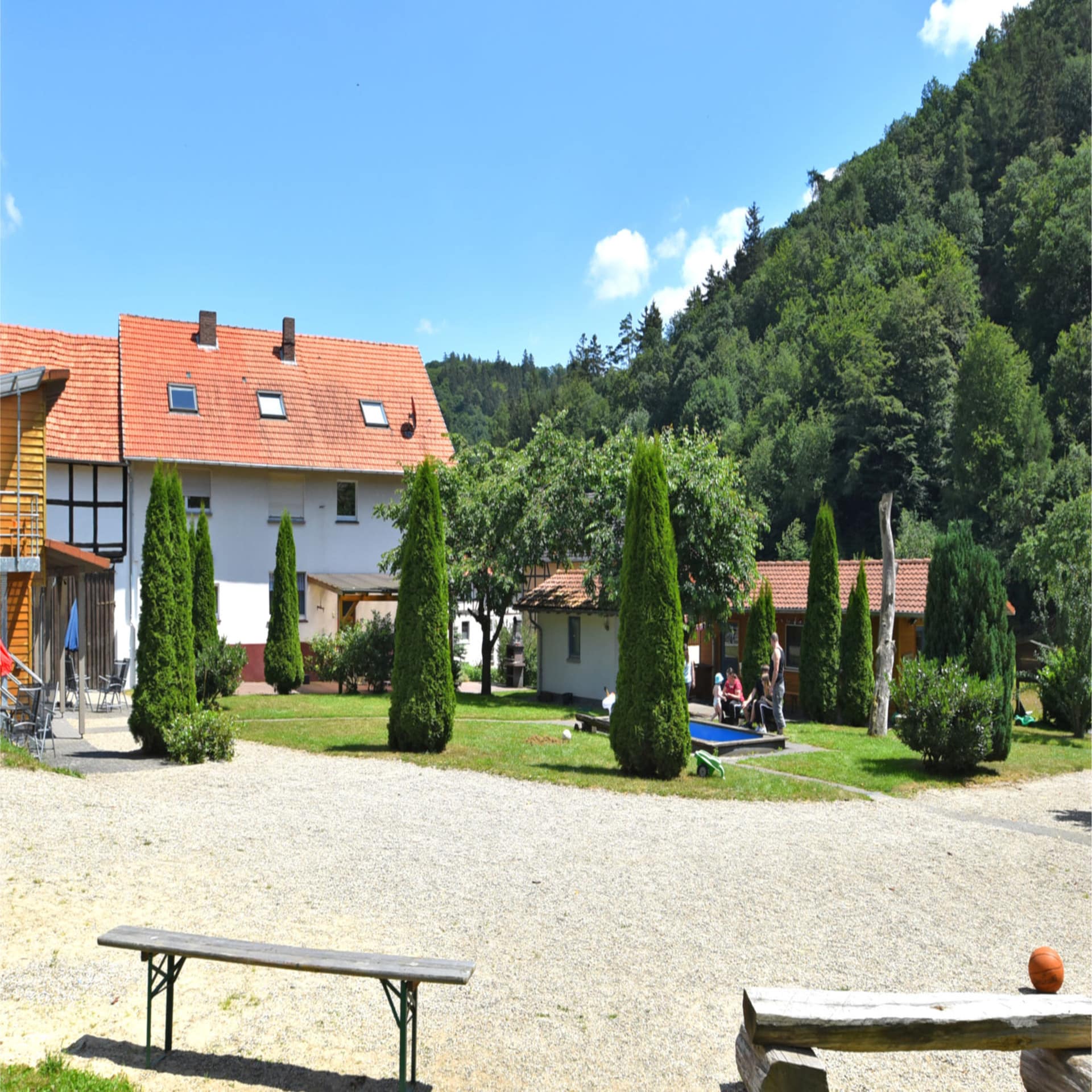 Weißes Bauernhaus mit Vorplatz auf dem sich ein kleiner Pool befindet. Umgeben von Bäumen. 