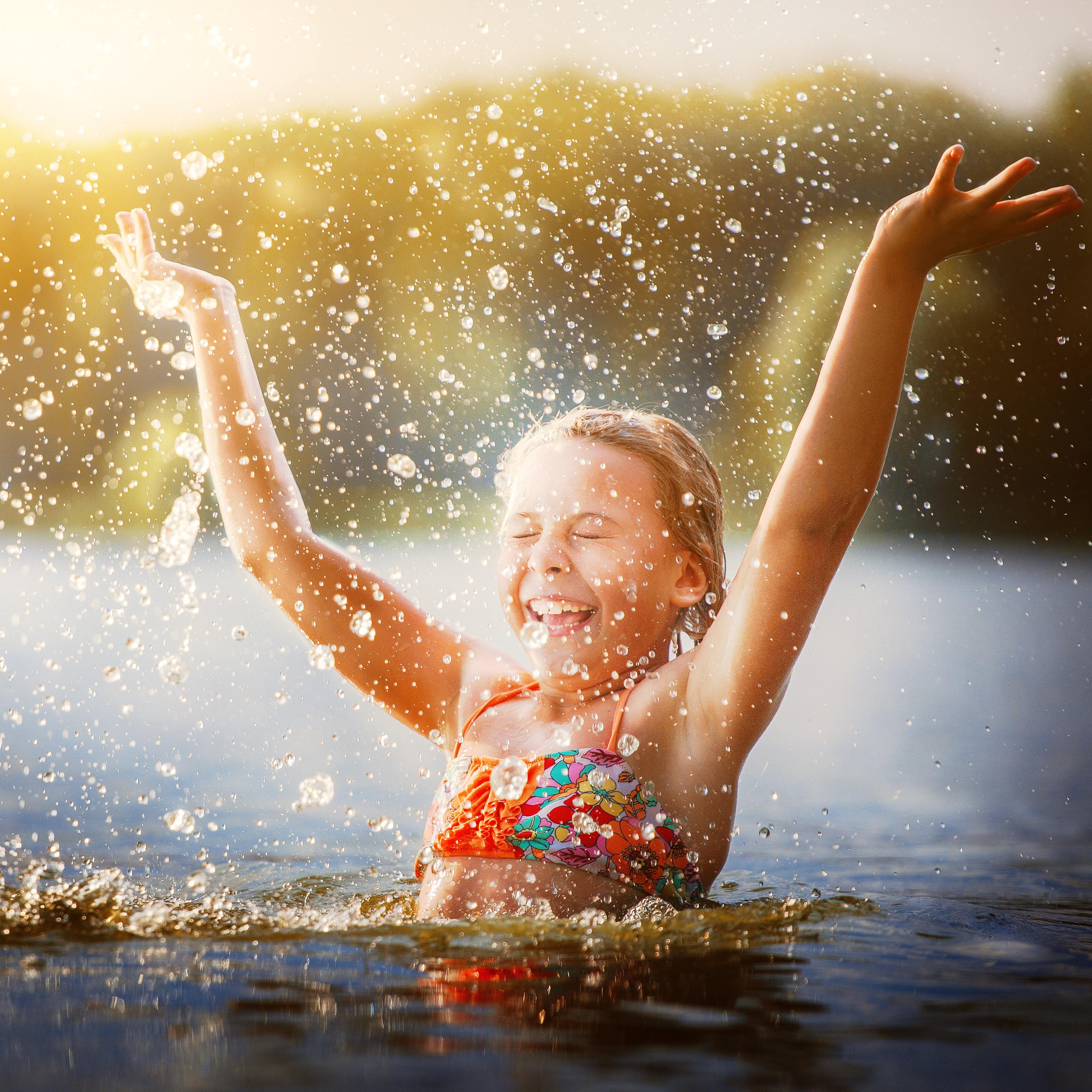 Ein Mädchen im Bikini steht bis über den Bauch im Wasser und spritzt mit geschlossenen Augen Wasser. 