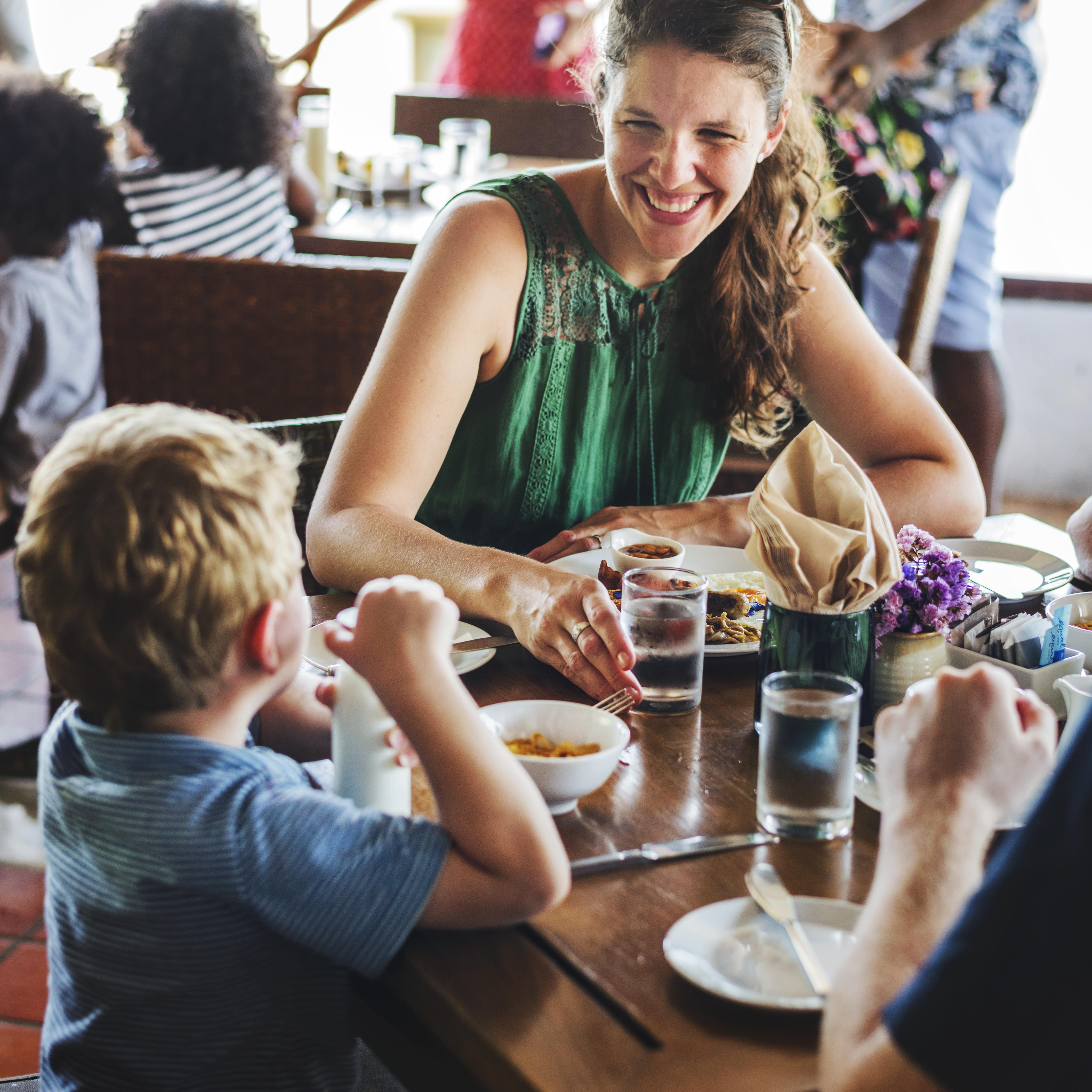 Familie mit 2 Kindern sitzt an einem hölzernen Frühstückstisch im Hotelrestaurant. Die Mutter lacht den Jungen an.