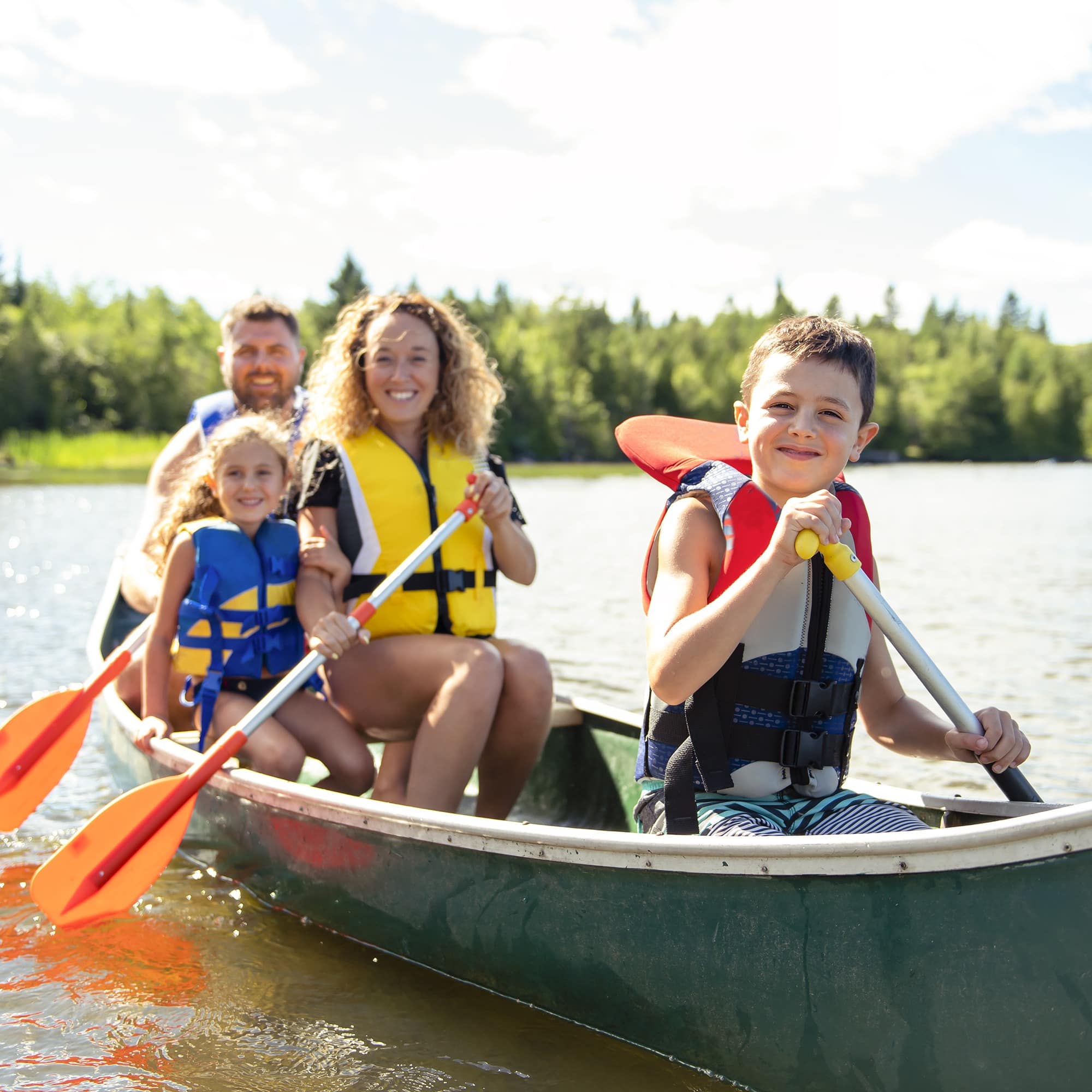 Familie mit 2 Kindern rudert in einem grünen Kanu auf einem See. Alle haben Schwimmwesten an.