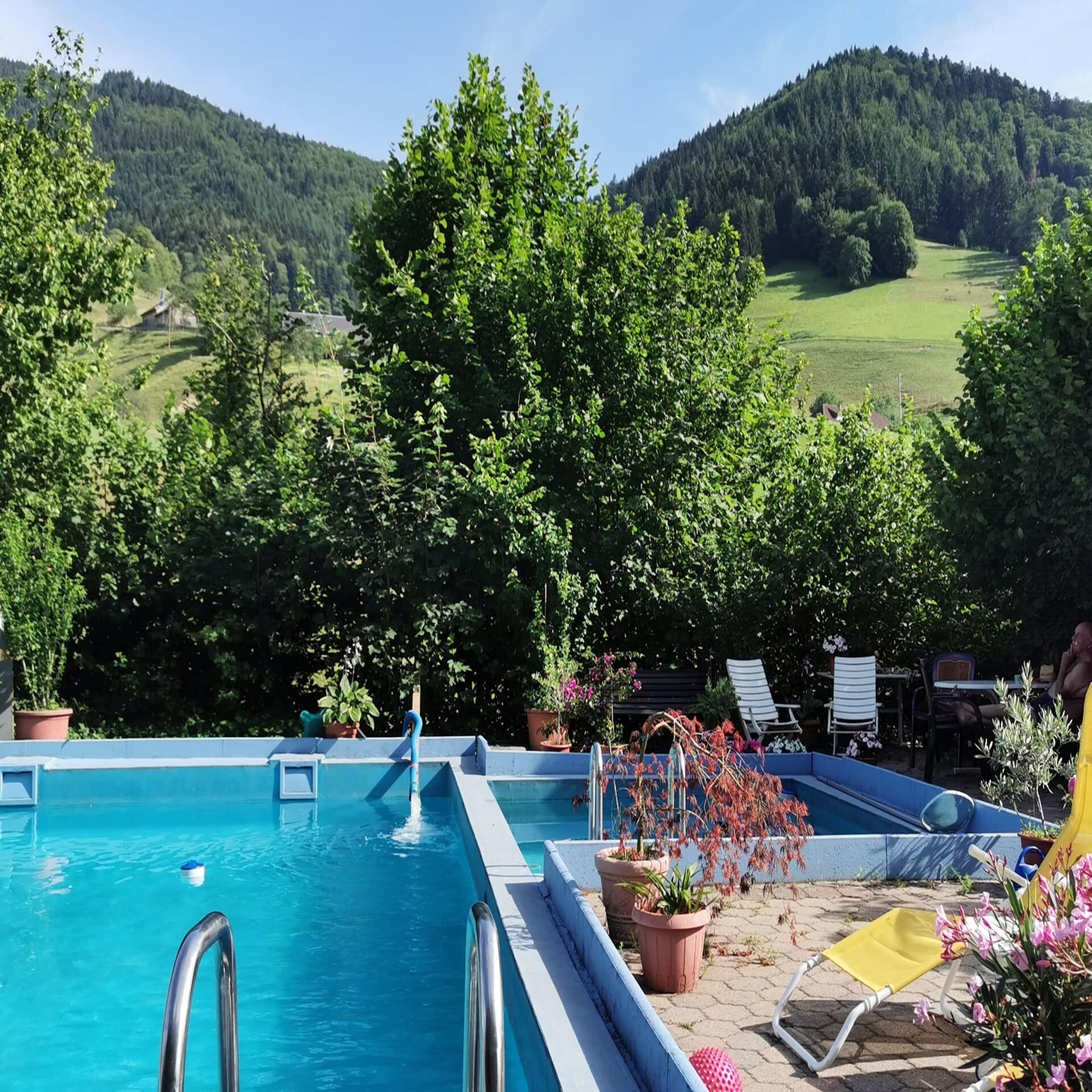 Blick über einen Garten mit Pool, dahinter das hügelige Umland im Sommer.