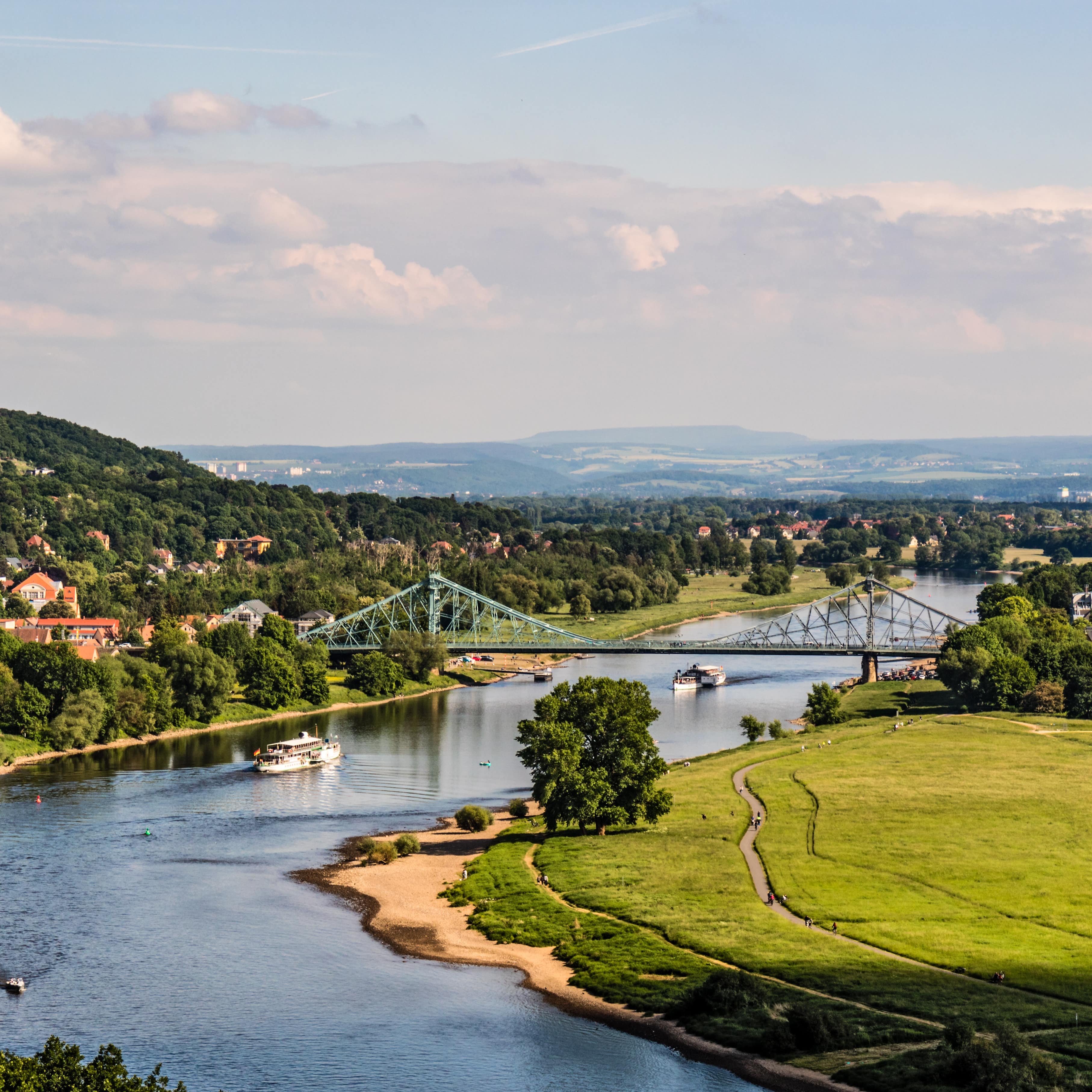 Blick über die Elbe und eine Brücke, rechts ein kleiner Strand und ein Spazierweg. Links Bäume und Häuser.