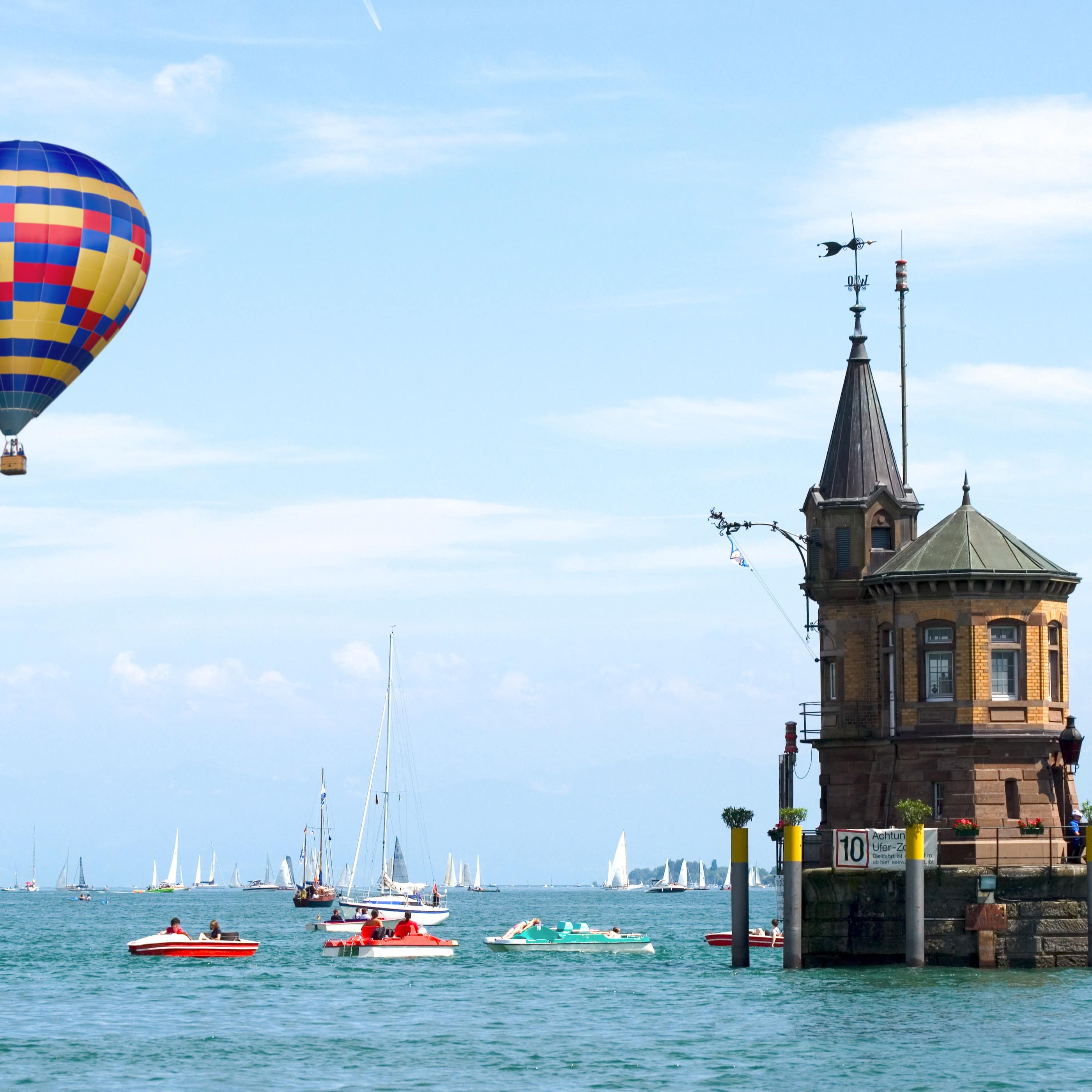 Ein bunter Heißluftballon fährt über dem Bodensee. Boote im Wasser, rechts der Hafen von Konstanz.