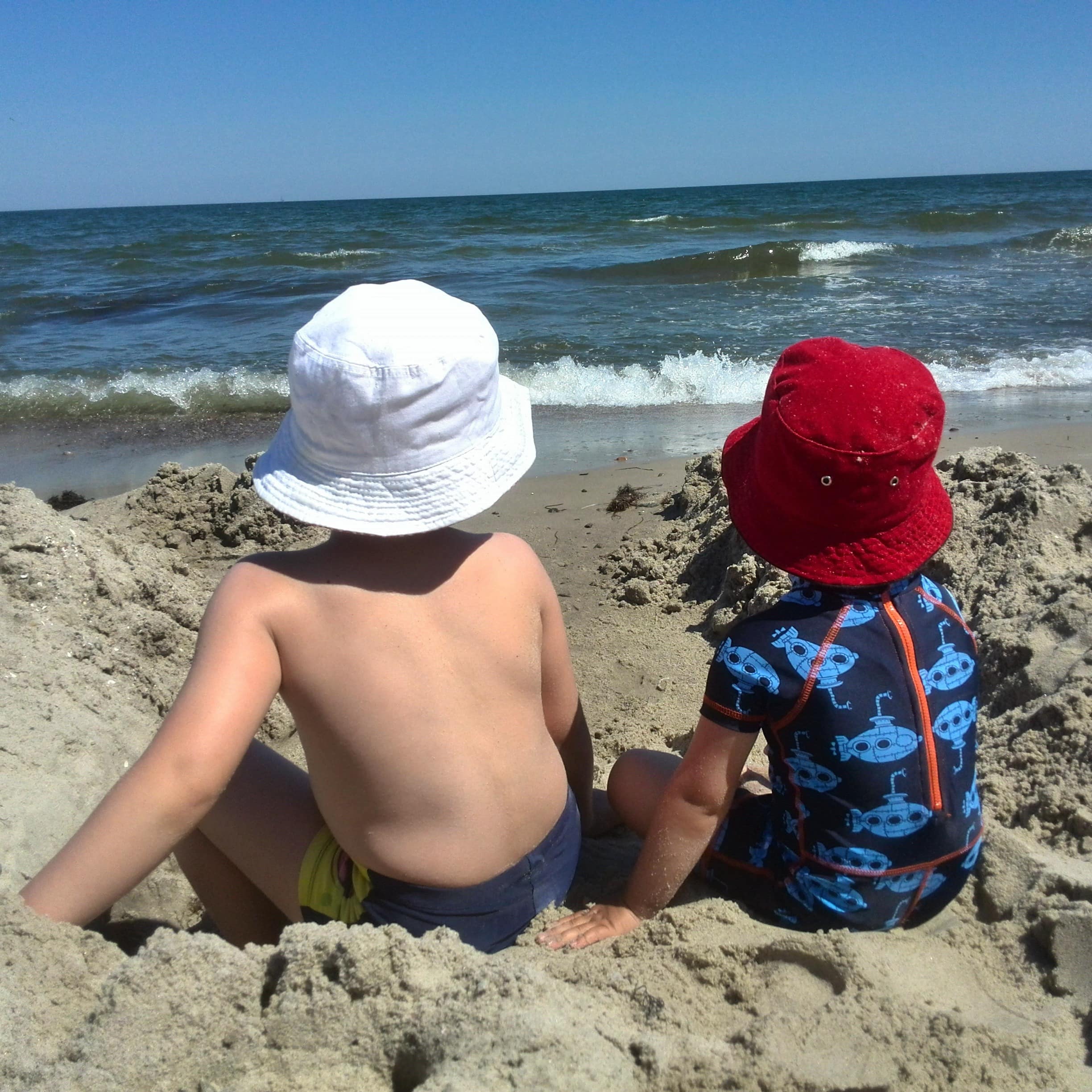Rückenansicht: 2 Jungen sitzen im Sand direkt am Wasser. 