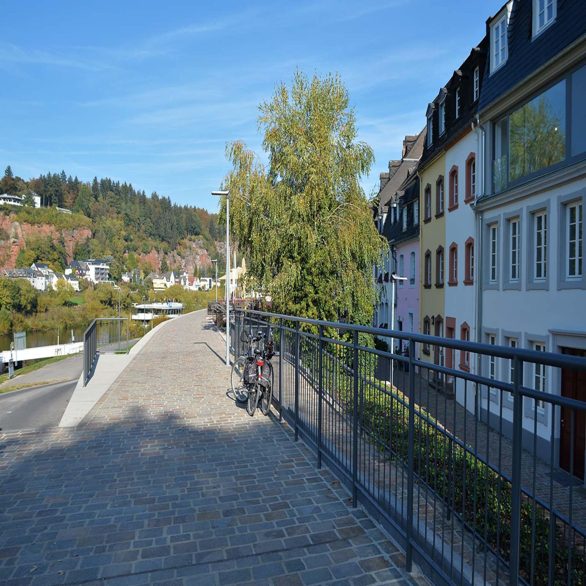 Promenade am Flussufer, rechts Häuser, davor ein Geländer, an dem 2 Fahrräder lehnen.