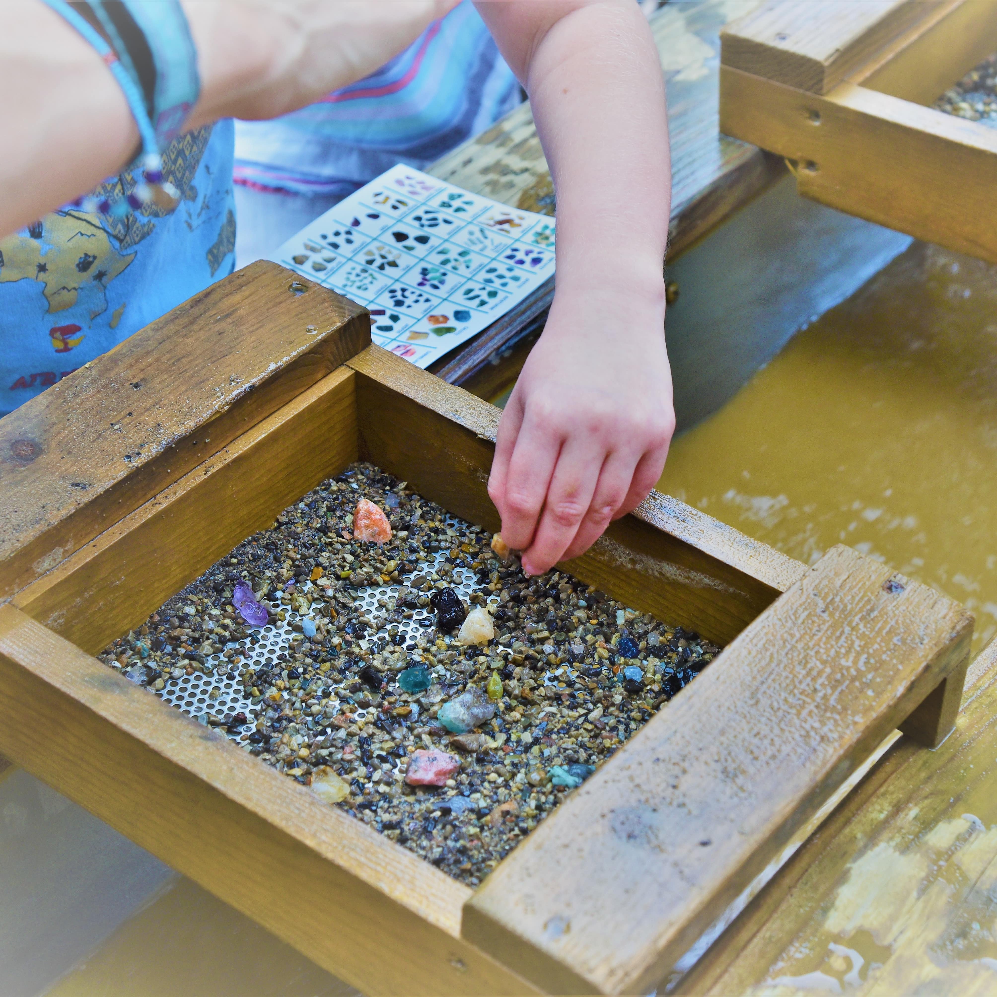 Nahaufnahme: Eine Kinderhand siebt Edelsteine in einem Holzsieb über Wasser.