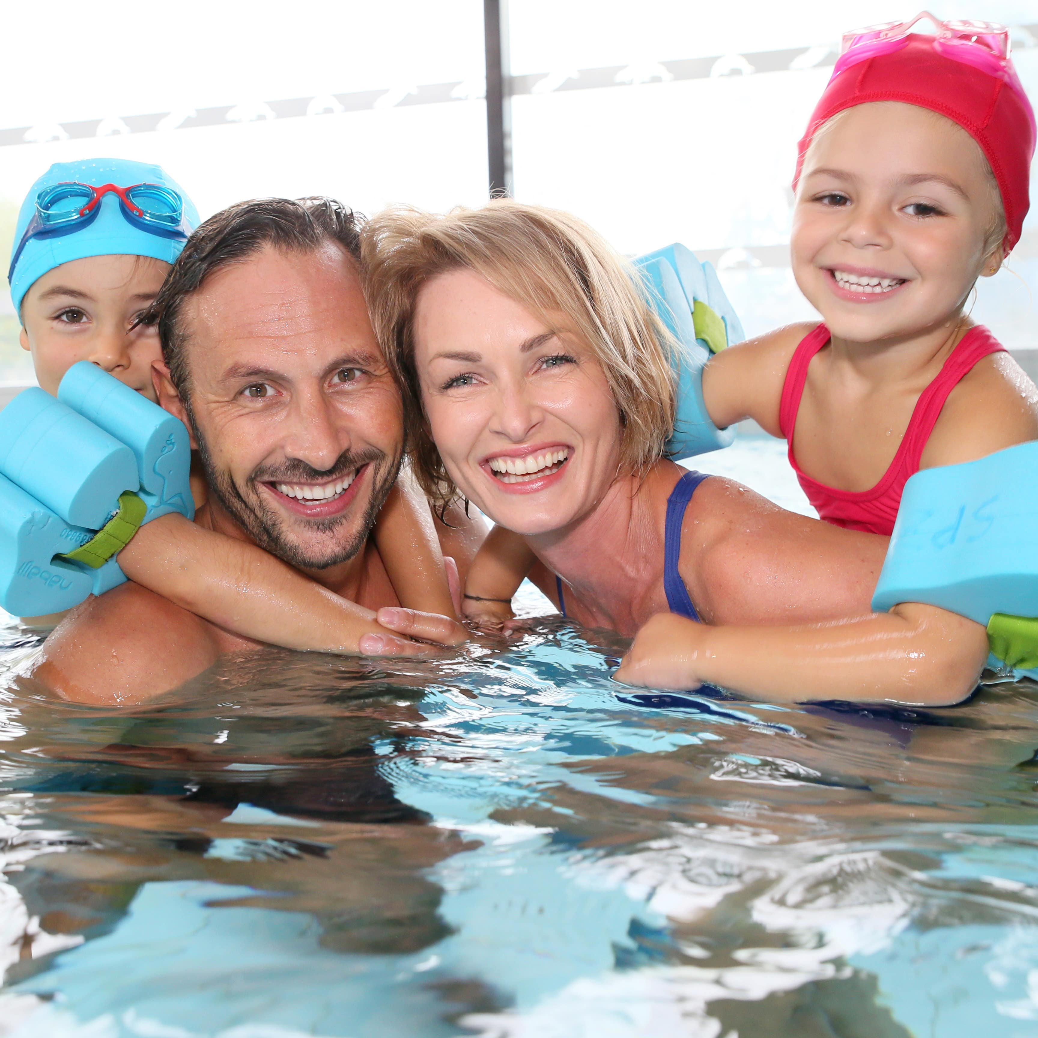 Eltern mit 2 Kindern vergnügen sich in einem Indoor-Pool und lachen in die Kamera. Die Kinder haben Badekappen auf.