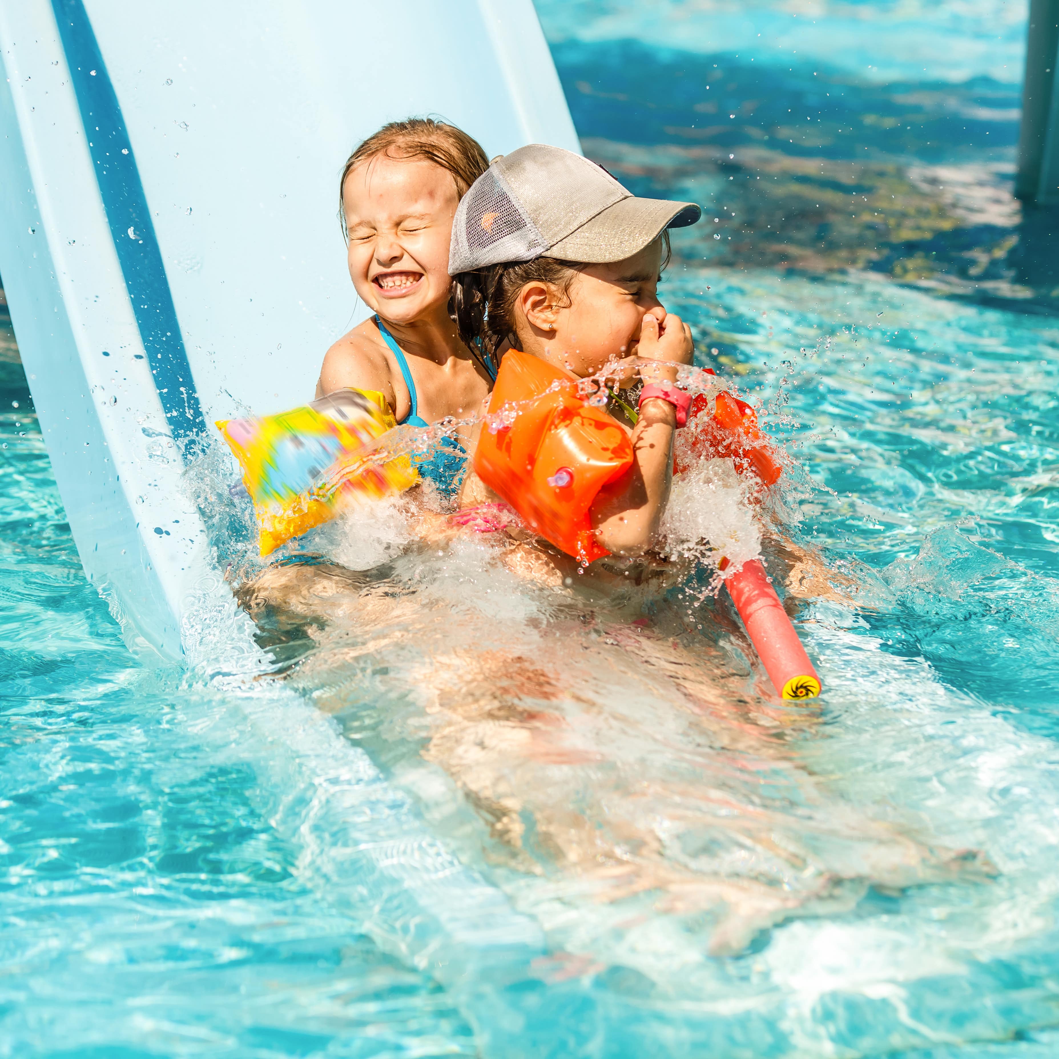 2 kleine Kinder mit Schwimmflügeln auf einer Wasserrutsche. Beide haben die Augen zu, eines hält sich die Nase zu.