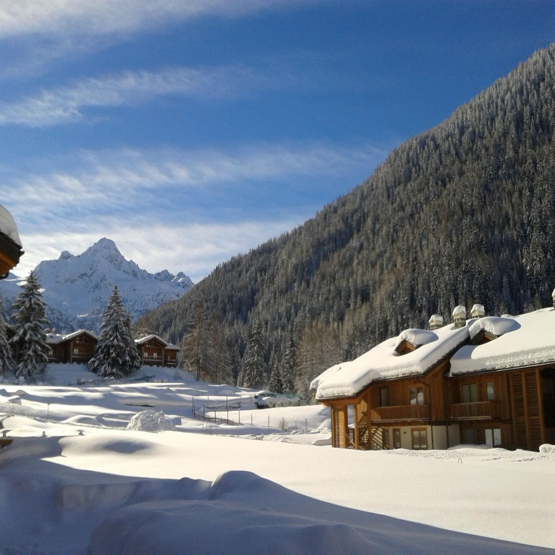 Ein paar verschneite Hütten im Skigebiet La Thuile. Rechts Bäume, im Hintergrund Berge. Die Sonne scheint.