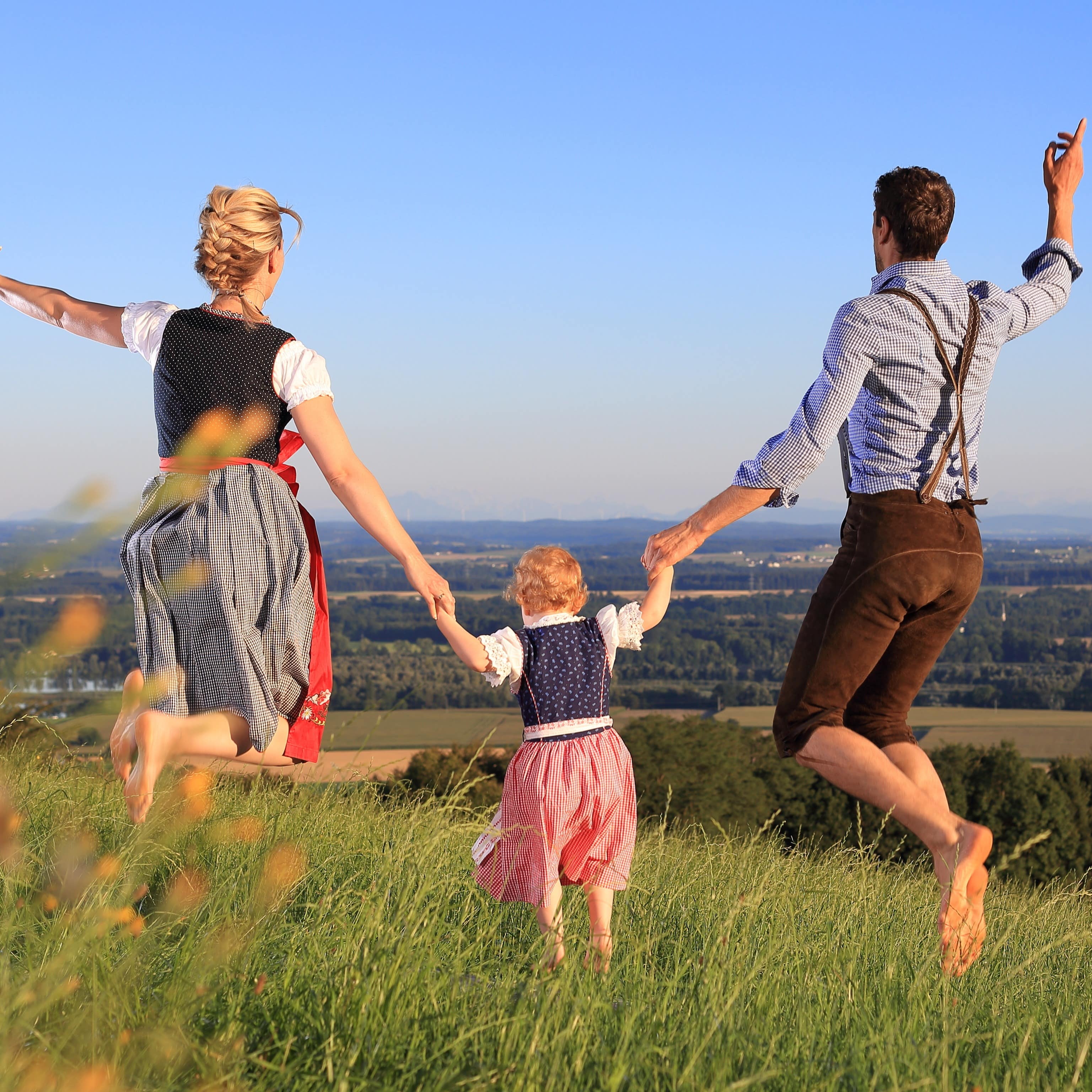 Rückenansicht: Eltern und ein kleines Mädchen in Tracht springen auf einem Hügel in die Luft. 